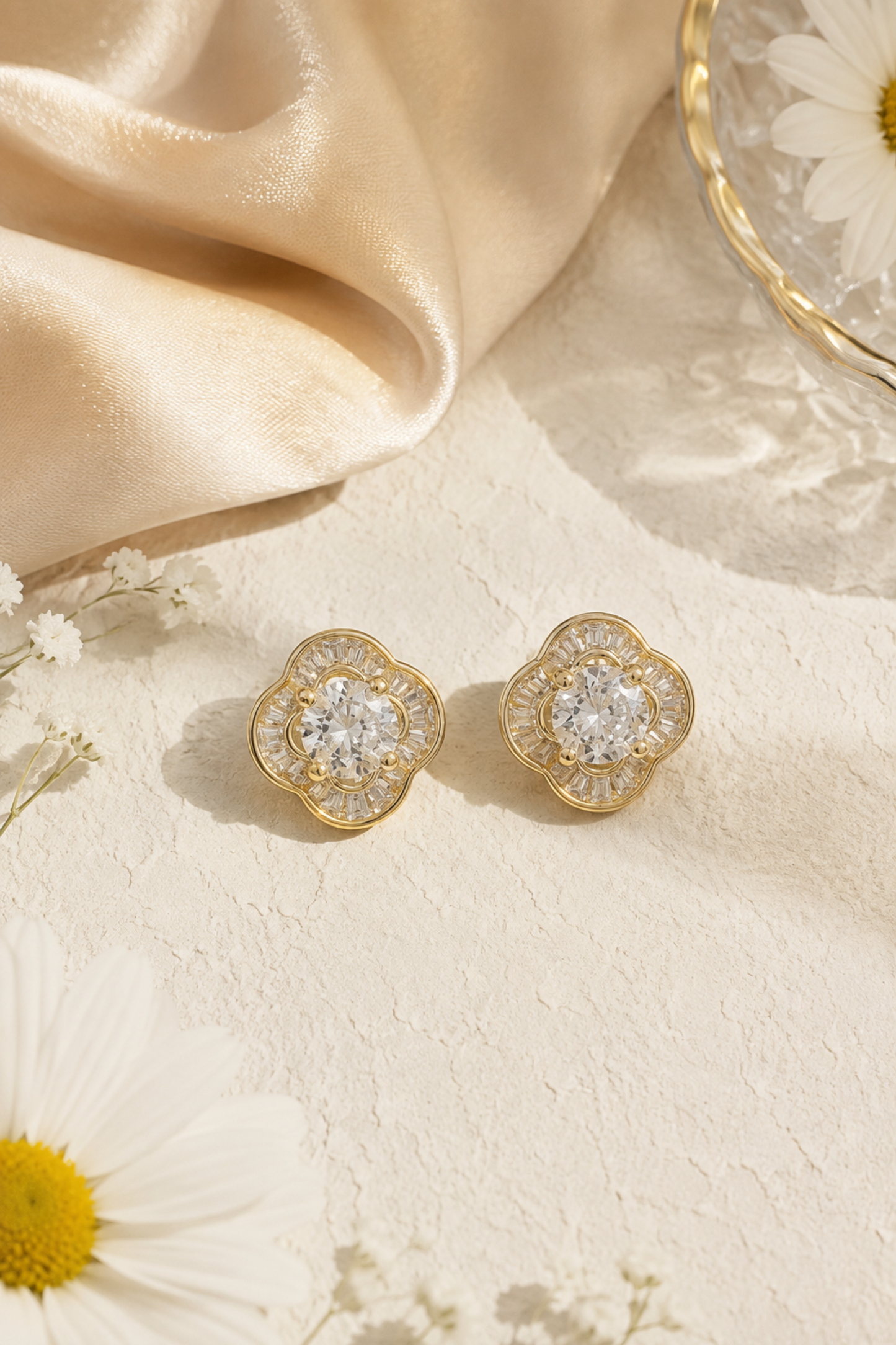 A pair of gold-toned flower-shaped stud earrings with clear gemstones, arranged on a textured cream surface with draped satin fabric and daisy flowers as styling props.