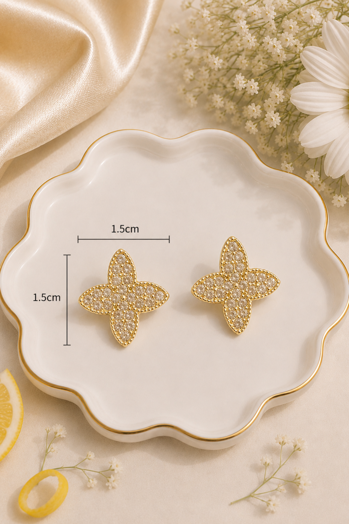 A pair of gold-toned flower-shaped stud earrings with clear rhinestone-like embellishments, displayed on a scalloped white tray with measurement markers showing 1.5cm by 1.