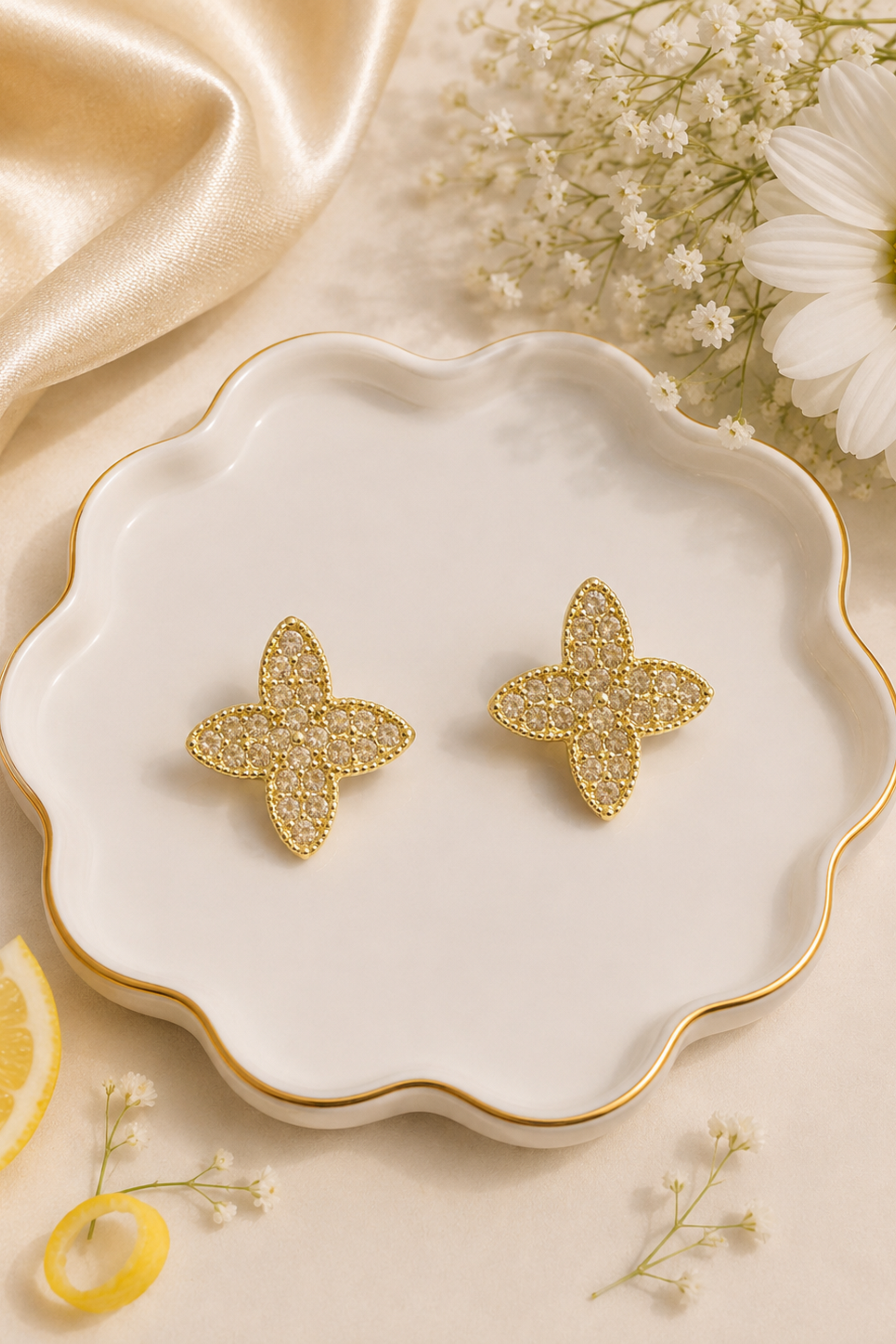 A pair of gold-toned flower-shaped stud earrings with textured pavé detailing, displayed on a scalloped white ceramic dish with gold trim, surrounded by decorative flowers, a lemon slice, and gold satin fabric.