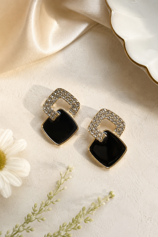 A pair of gold-toned geometric drop earrings with black square-shaped stones and clear rhinestone-encrusted arch tops, styled on a cream surface with a white flower and satin fabric.