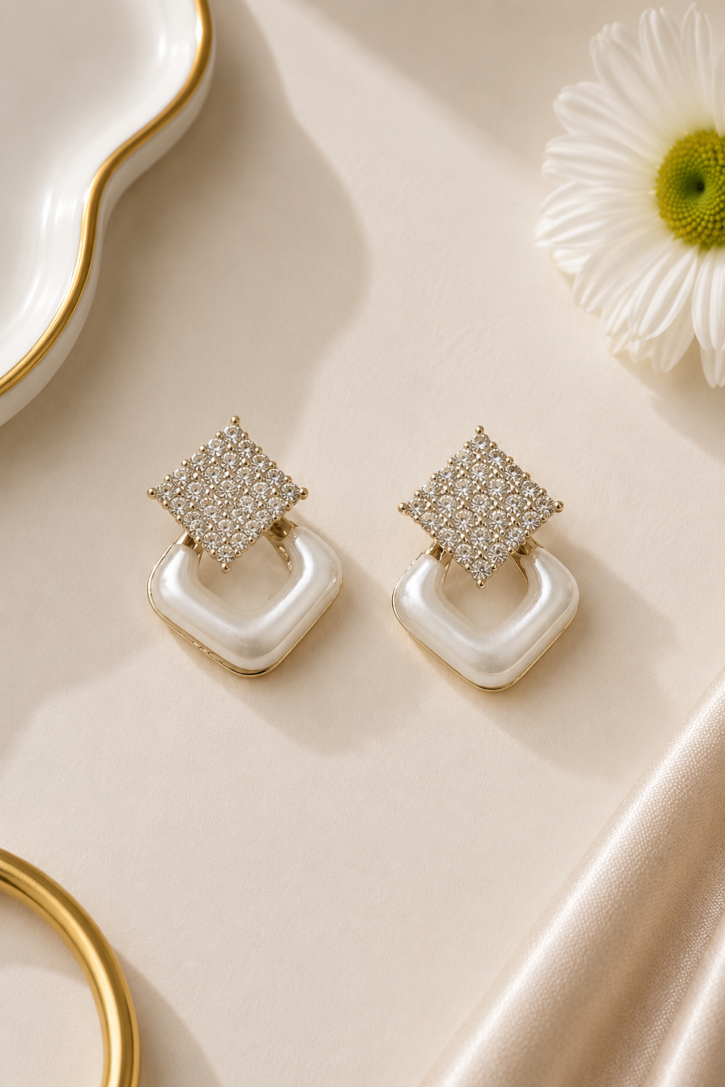 A pair of gold-toned geometric drop earrings with pavé-set clear stones on the top square and smooth white curved drop pendants, styled on a cream satin surface with a white flower and gold-rim dish props.