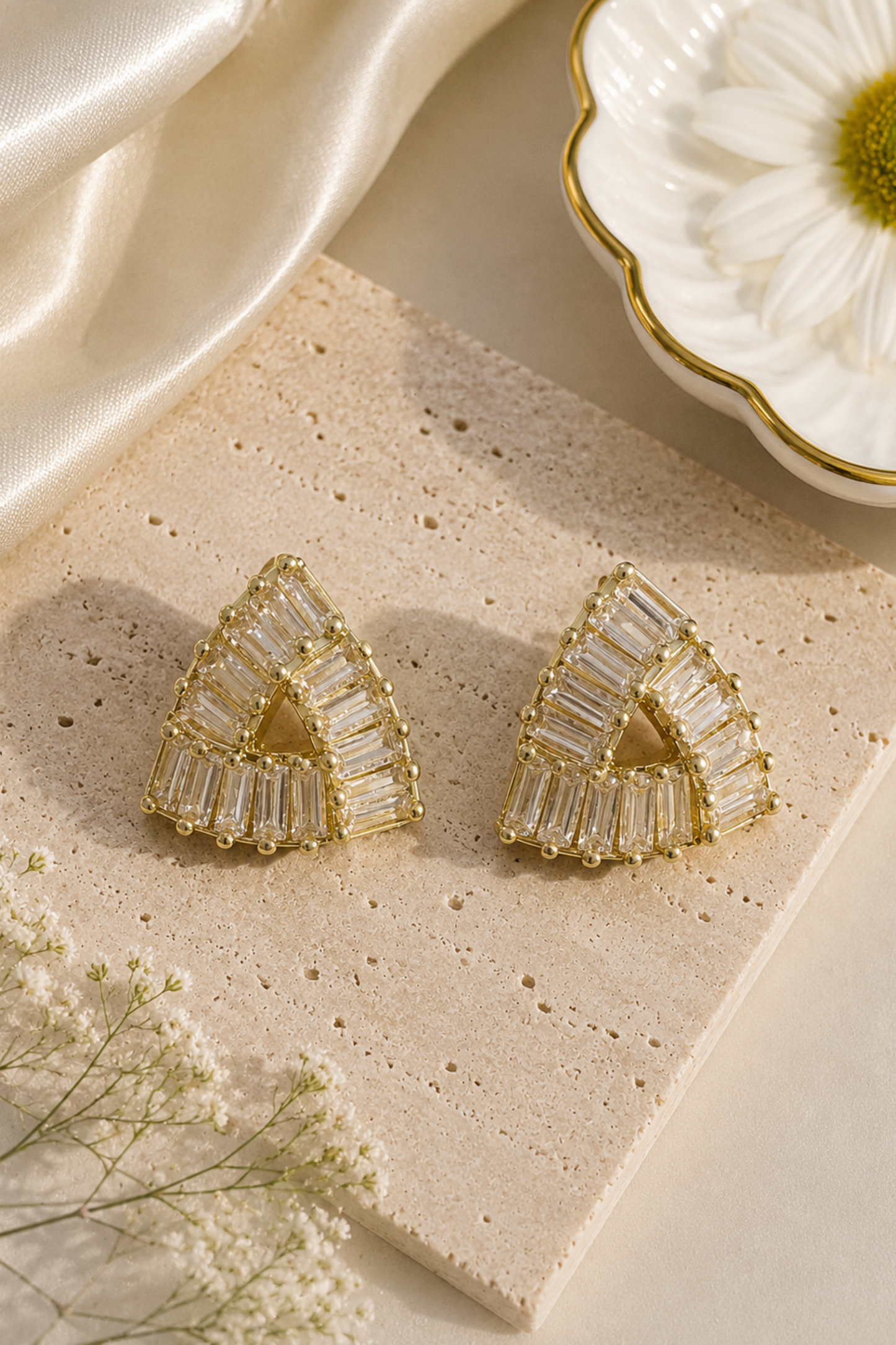 A pair of gold-toned geometric stud earrings with clear rectangular baguette stones, arranged on a beige stone slab with cream satin fabric, a white flower, and small white blossoms.
