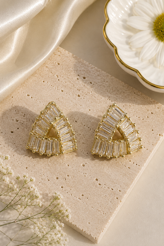 A pair of gold-toned geometric stud earrings with clear rectangular baguette stones, arranged on a beige stone slab with cream satin fabric, a white flower, and small white blossoms.