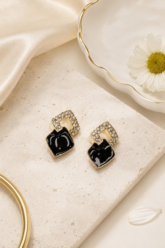 A pair of gold-toned geometric stud earrings with glossy black square drops and clear crystal accents, styled on a beige stone slab with satin fabric, a white daisy, and a gold hoop in the background.
