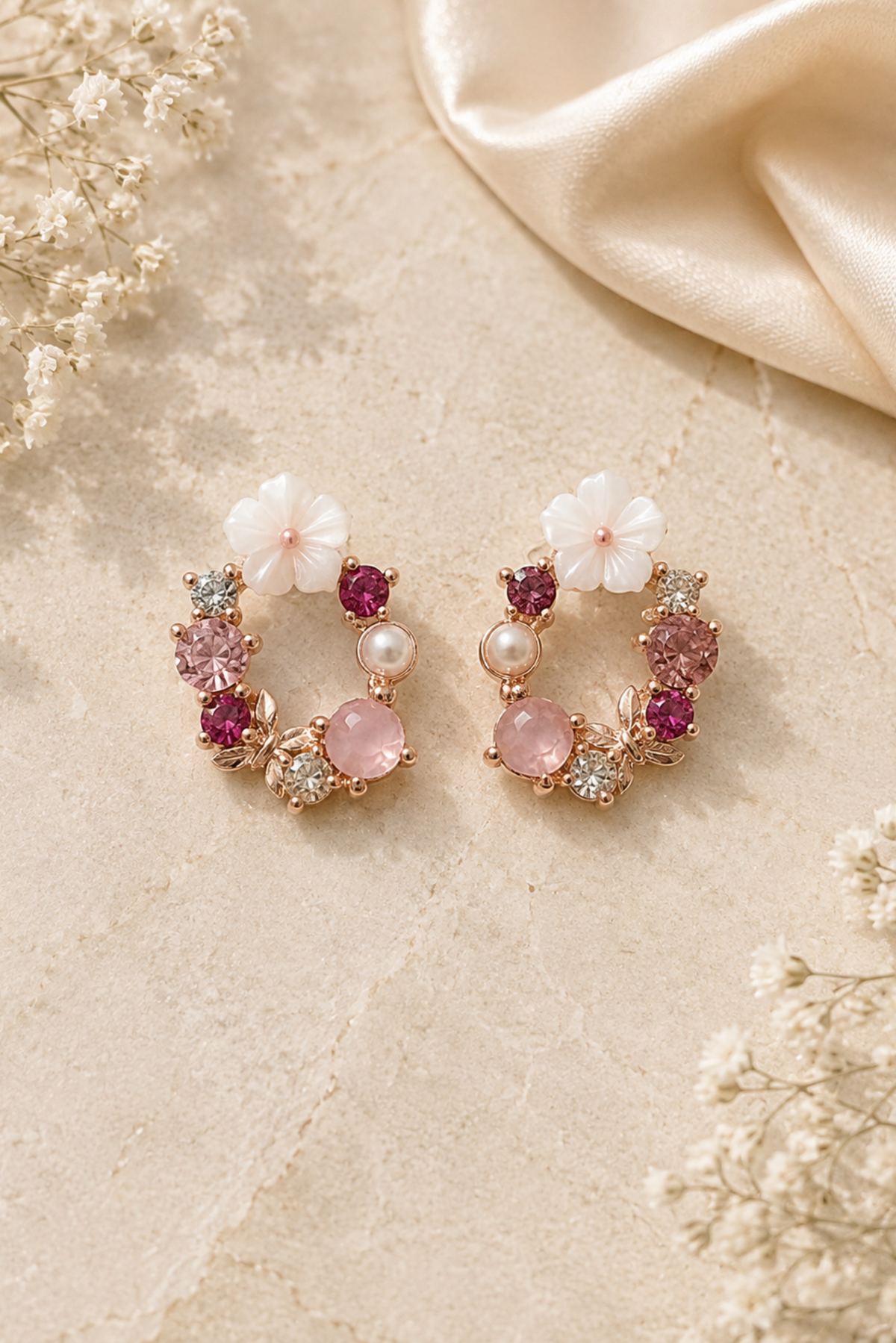 A pair of gold-toned hoop earrings decorated with white flower charms, pink gemstones, and small pearl accents, displayed on a textured beige surface with dried flowers and a satin fabric.
