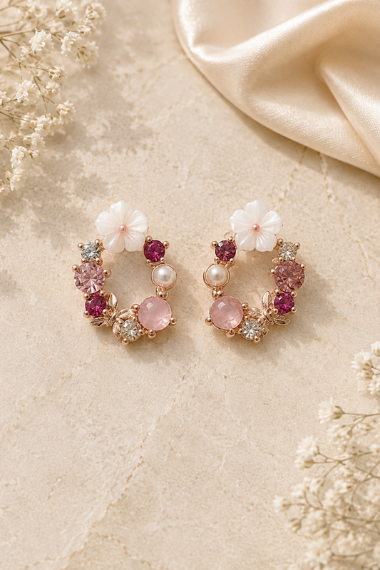A pair of gold-toned hoop earrings decorated with white flower charms, pink gemstones, and small pearl accents, displayed on a textured beige surface with dried flowers and a satin fabric.