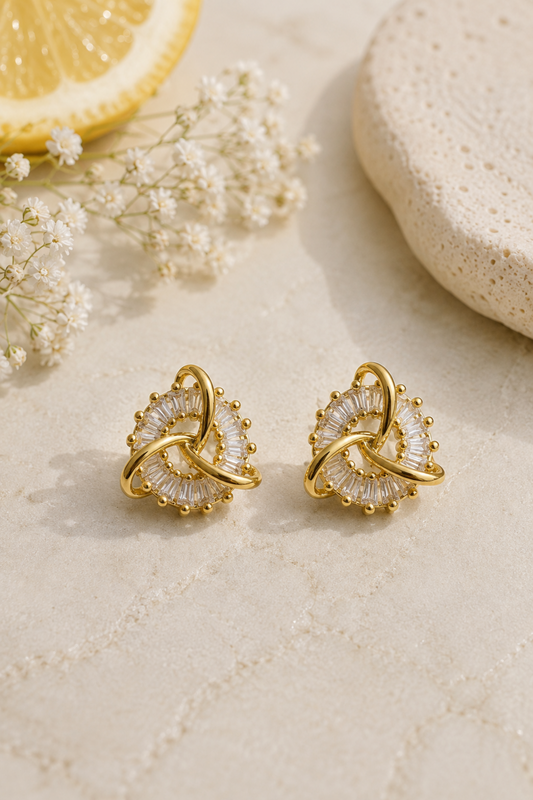 A pair of gold-toned hoop earrings with clear faceted crystal-like accents, styled on a light beige textured surface with a lemon slice, white baby's breath flowers, and a stone prop.