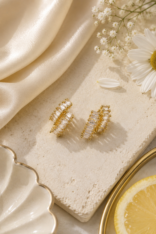A pair of gold-toned hoop earrings with clear rectangular crystals, styled on a textured cream stone with cream satin fabric, white flowers, and a lemon slice.