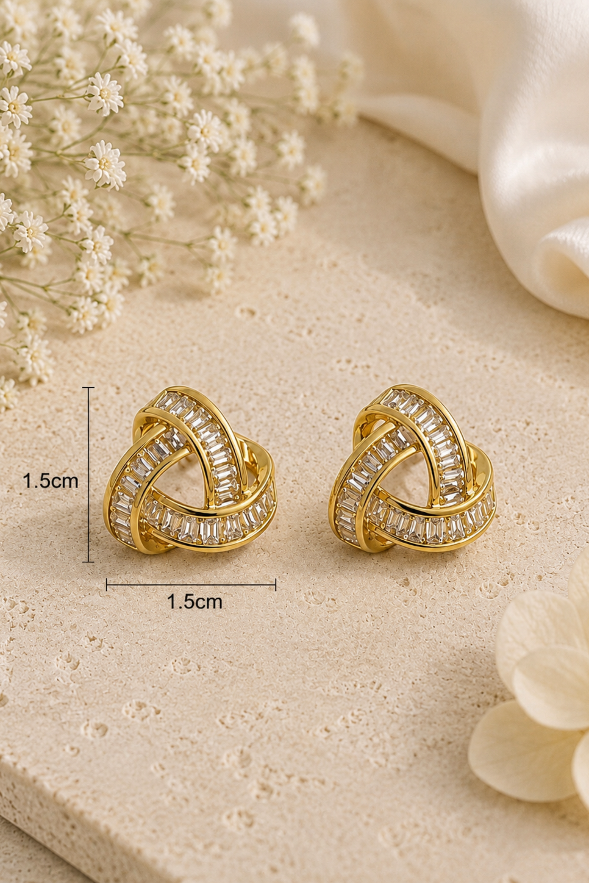 A pair of gold-toned knot-style hoop earrings with clear crystal-like stones, shown on a light beige textured surface with small white flowers and a white fabric prop. Black measurement lines and text indicate 1.