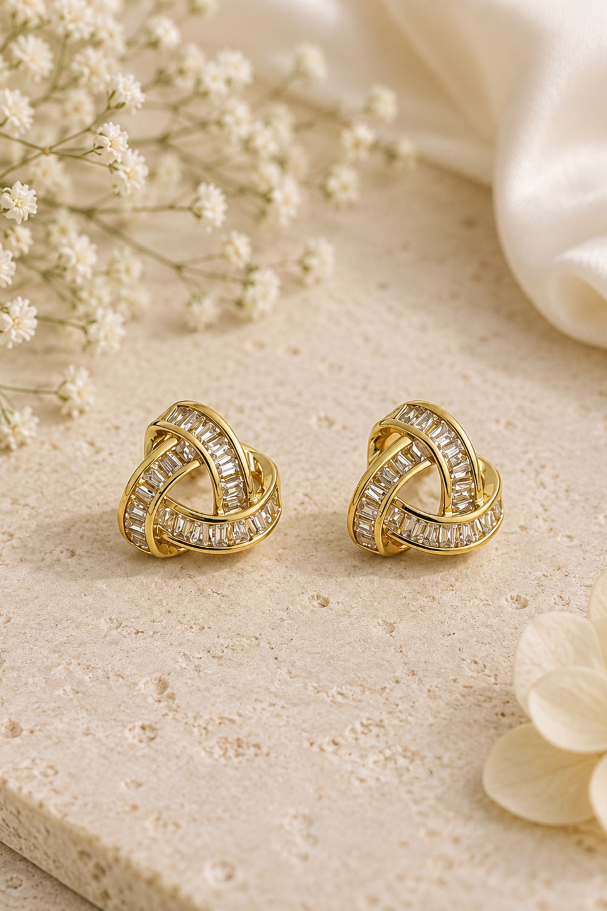 A pair of gold-toned knot-style stud earrings set with clear rectangular stones, displayed on a light beige textured surface with white baby's breath flowers and soft white fabric in the background.