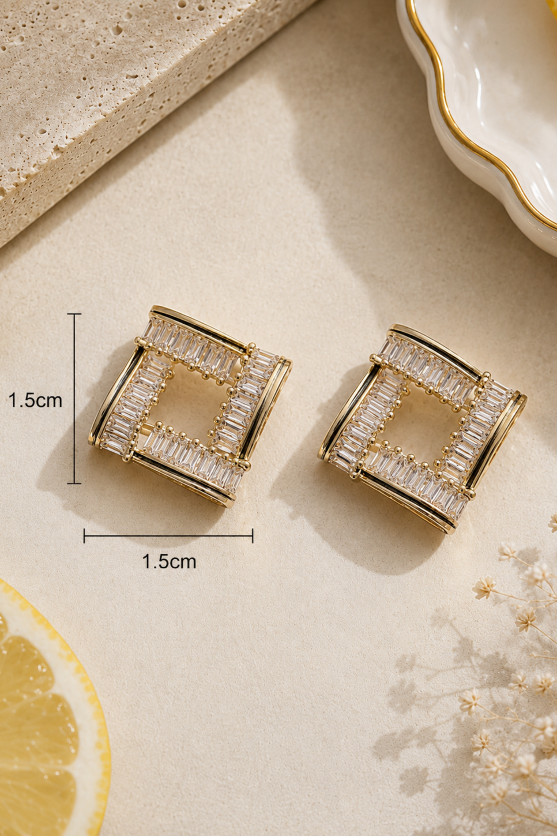 A pair of gold-toned square stud earrings with clear baguette crystals, shown on a beige surface with dimension markers indicating 1.5cm by 1.