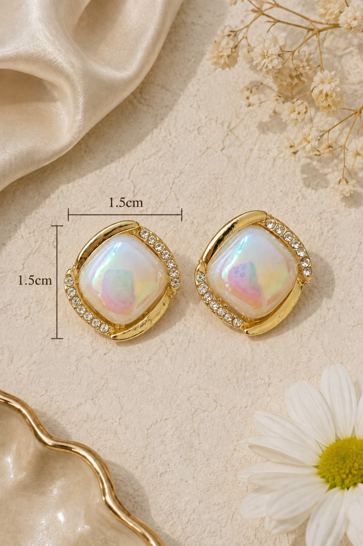 A pair of gold-toned square stud earrings with iridescent white center stones and clear crystal accents, shown on a beige textured surface with decorative fabric, dried flowers, and a white daisy. Measurement lines indicate 1.