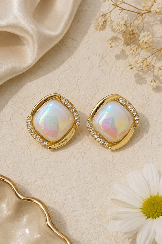 A pair of gold-toned square stud earrings with iridescent white center stones and small clear crystal accents, arranged on a textured cream surface with satin fabric, dried flowers, and a white daisy.