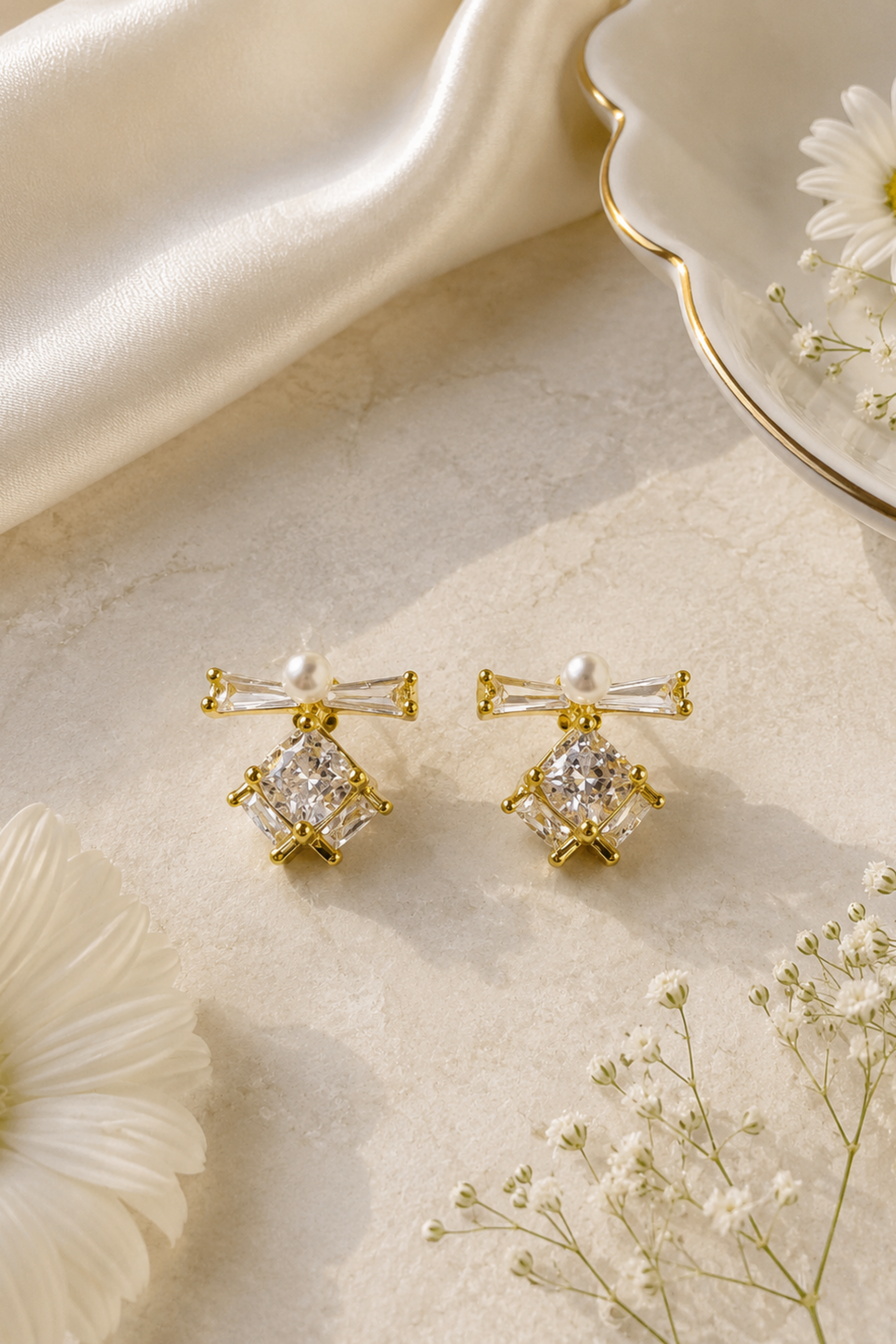 A pair of gold-toned statement earrings with clear crystal and pearl details, styled on a textured cream surface with white daisies and satin fabric.