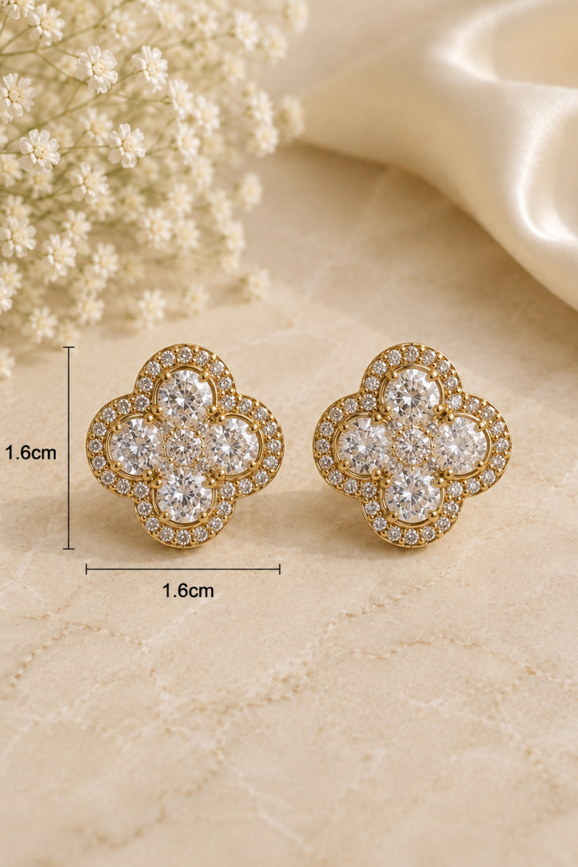 A pair of gold-toned stud earrings with a floral clover design set with clear stones, displayed on a beige textured surface with decorative baby's breath and satin fabric. Measurement lines and text indicating 1.