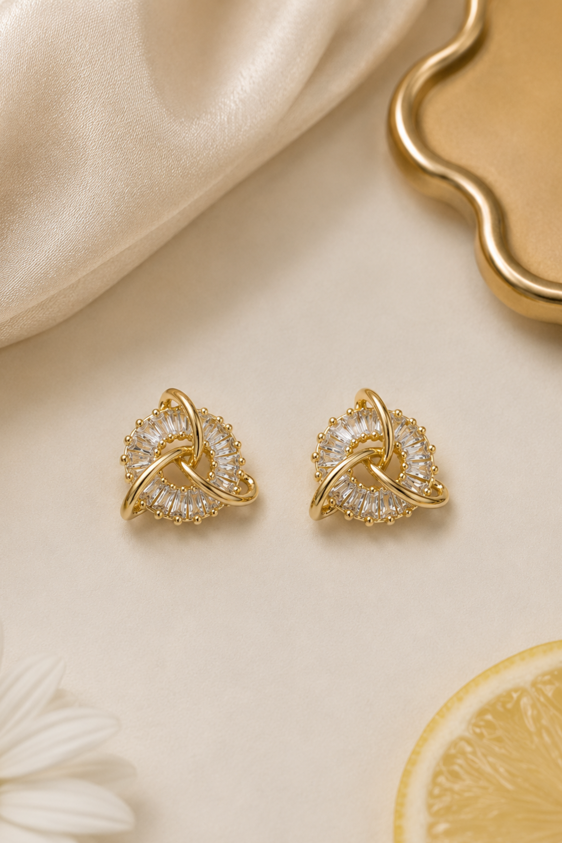 A pair of gold-toned stud earrings with a knot-like design and clear crystal-like accents, arranged on a warm cream surface with draped satin, a daisy flower, and a lemon slice.