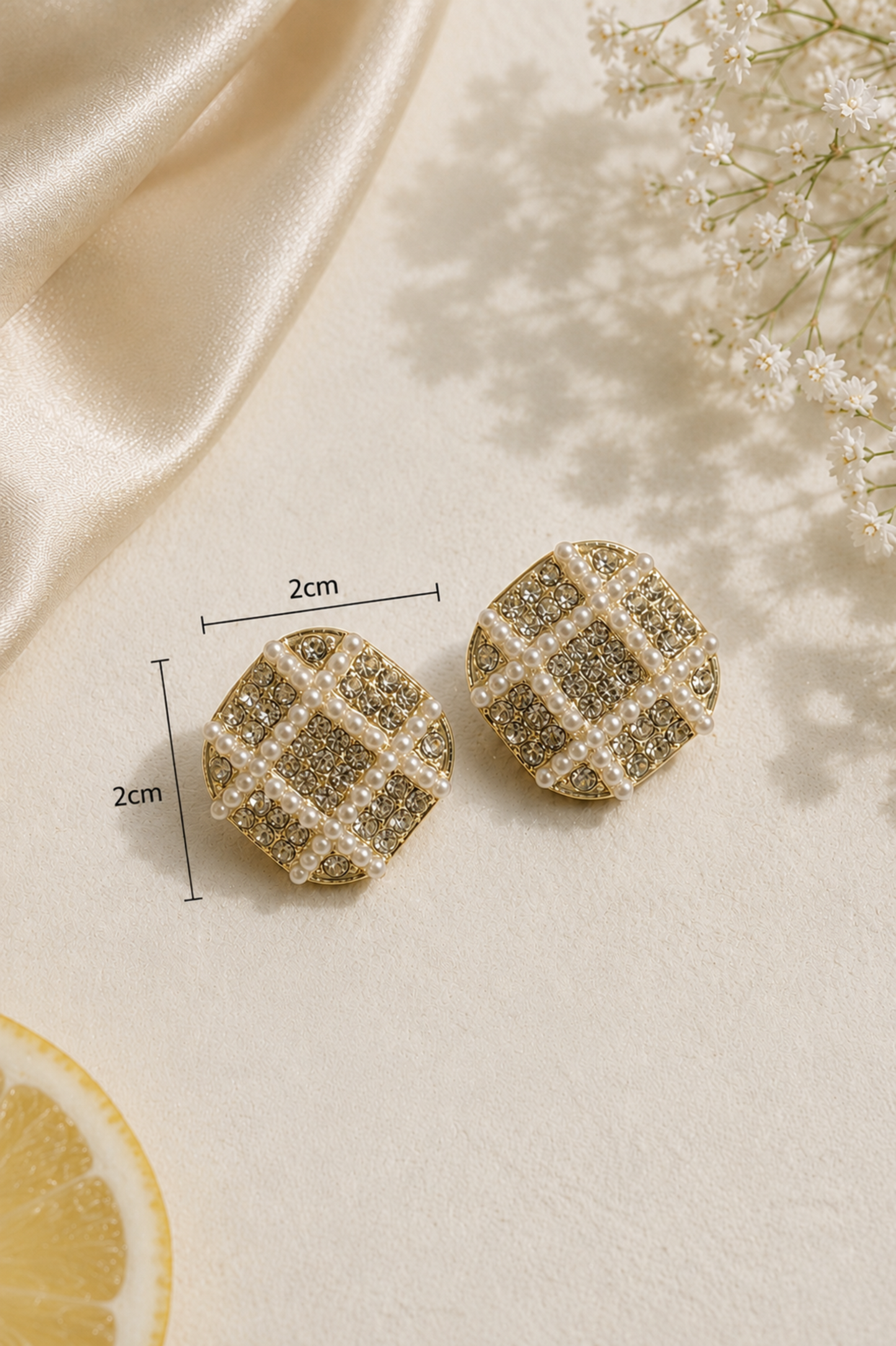 A pair of gold-toned stud earrings with a textured grid pattern of clear rhinestones and white faux pearls, displayed on a cream satin surface with decorative flowers, a lemon slice, and dimension lines indicating a size of 2cm by 2cm.