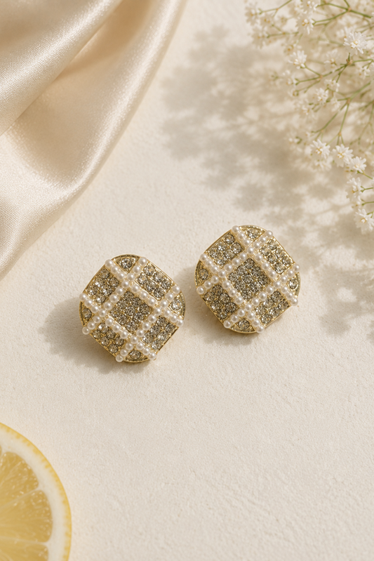 A pair of gold-toned stud earrings with a textured grid pattern of small clear stones, styled on a cream textured surface with cream satin fabric, baby's breath flowers, and a lemon slice.