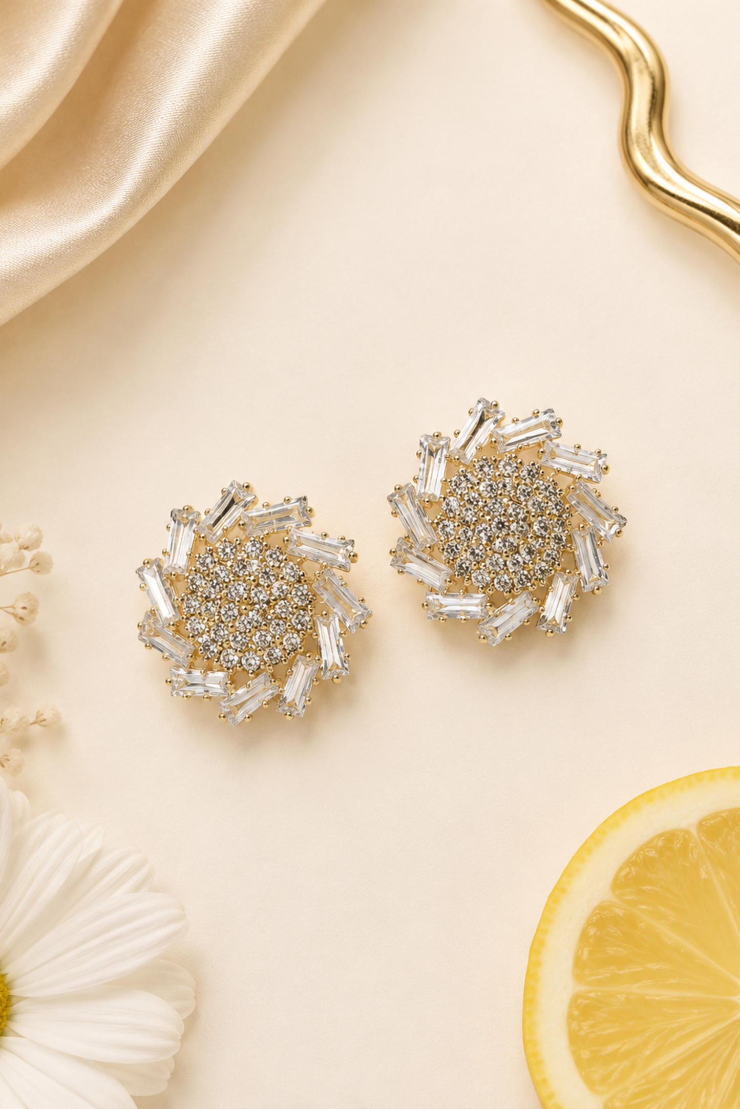 A pair of gold-toned stud earrings with clear crystal-like baguette and round stones, arranged on a warm cream background with satin fabric, a daisy flower, baby's breath, and a lemon slice.
