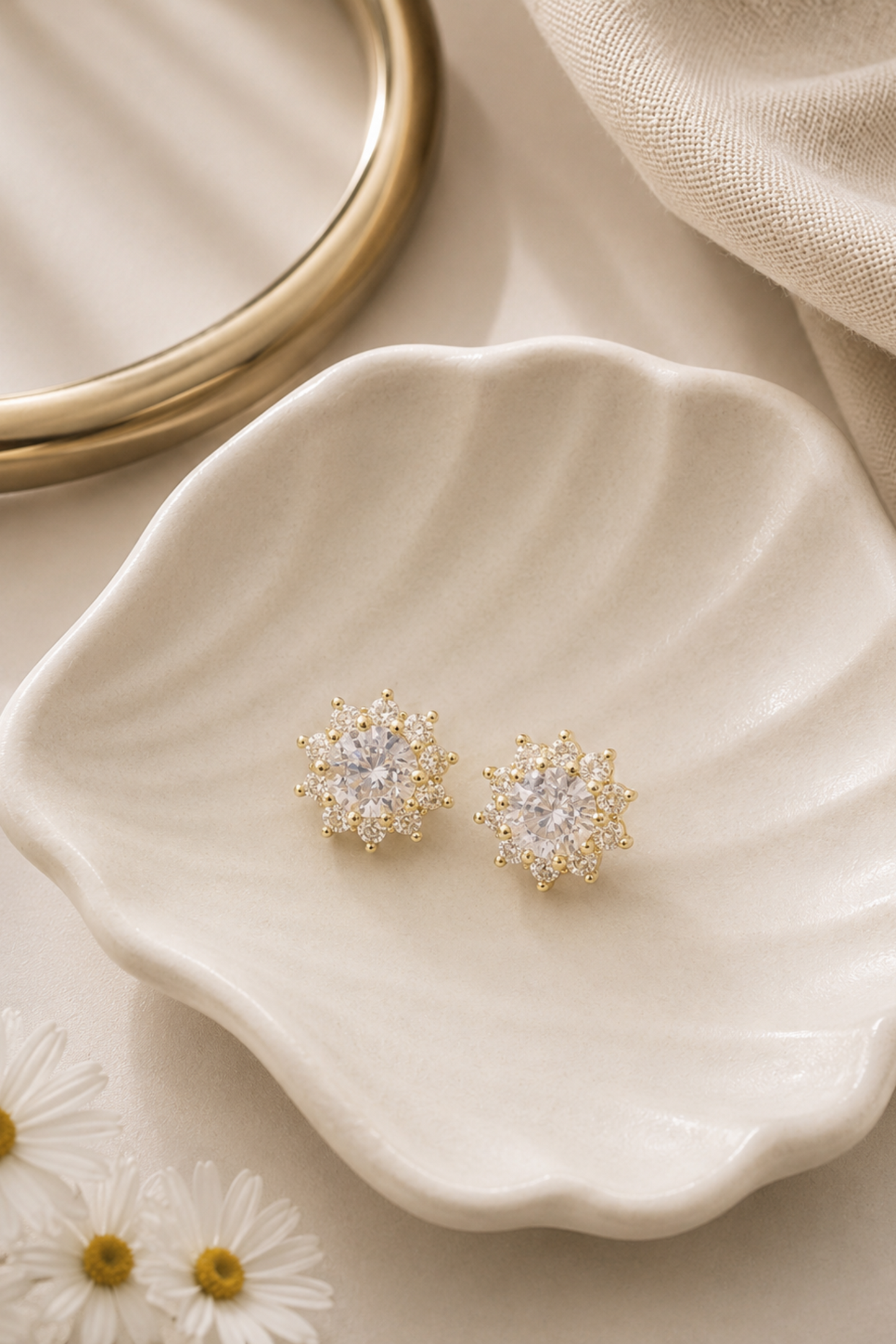 A pair of gold-toned stud earrings with clear crystal-like centers, displayed on a scalloped cream dish with daisies and a gold ring partially visible in a styled flat-lay setting.