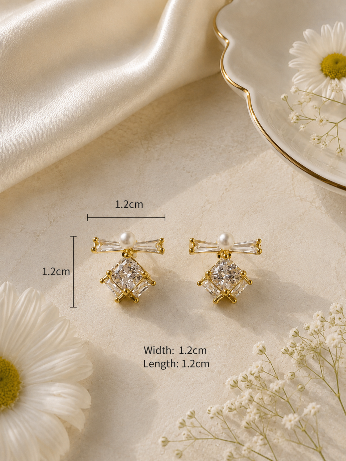 A pair of gold-toned stud earrings with clear gemstones and pearl-like accents, shown on a beige surface with decorative flowers and a plate. Measurement labels indicate 1.