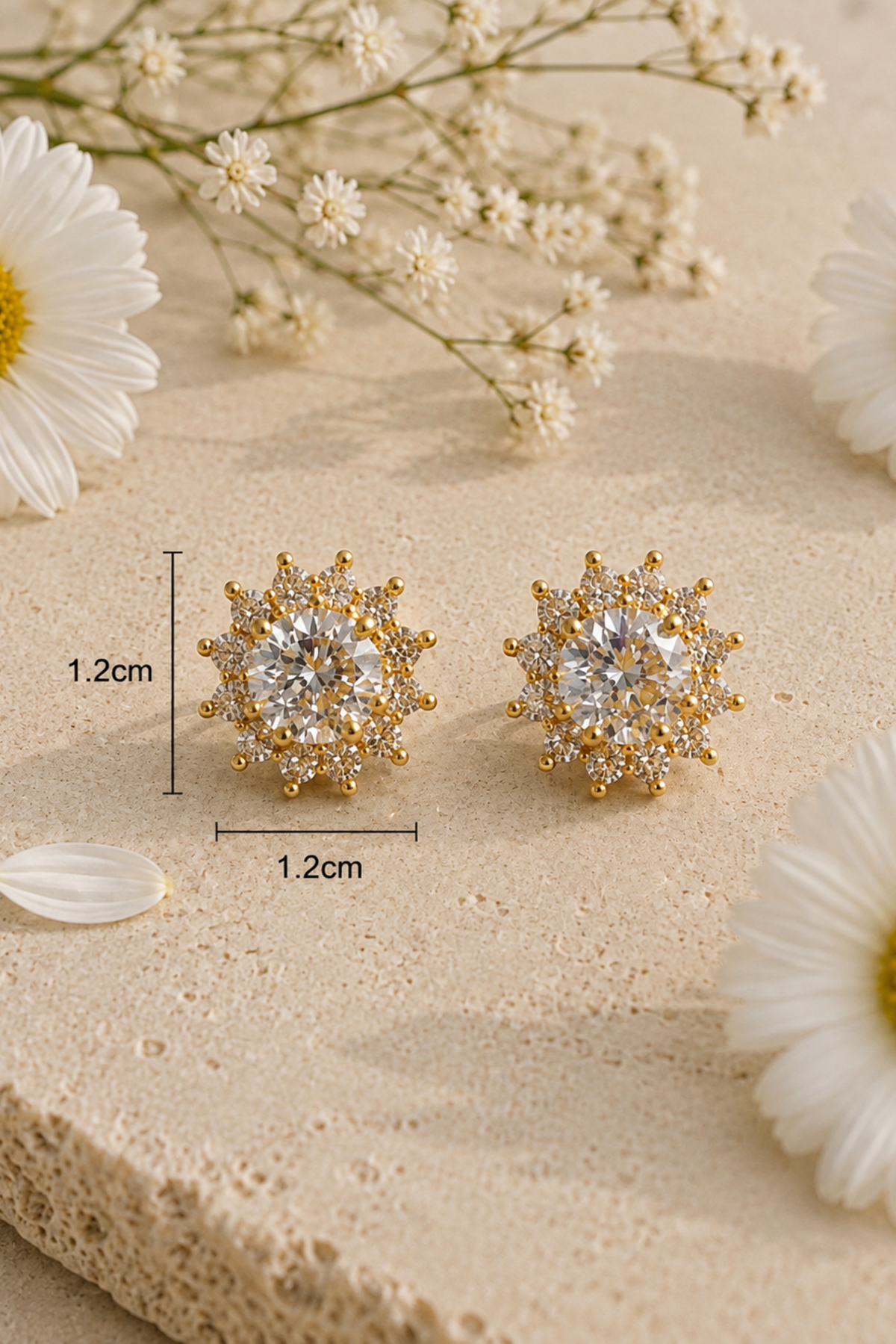 A pair of gold-toned stud earrings with clear gemstones arranged on a beige textured surface with white daisies and baby's breath flowers. Black measurement lines and text indicate 1.