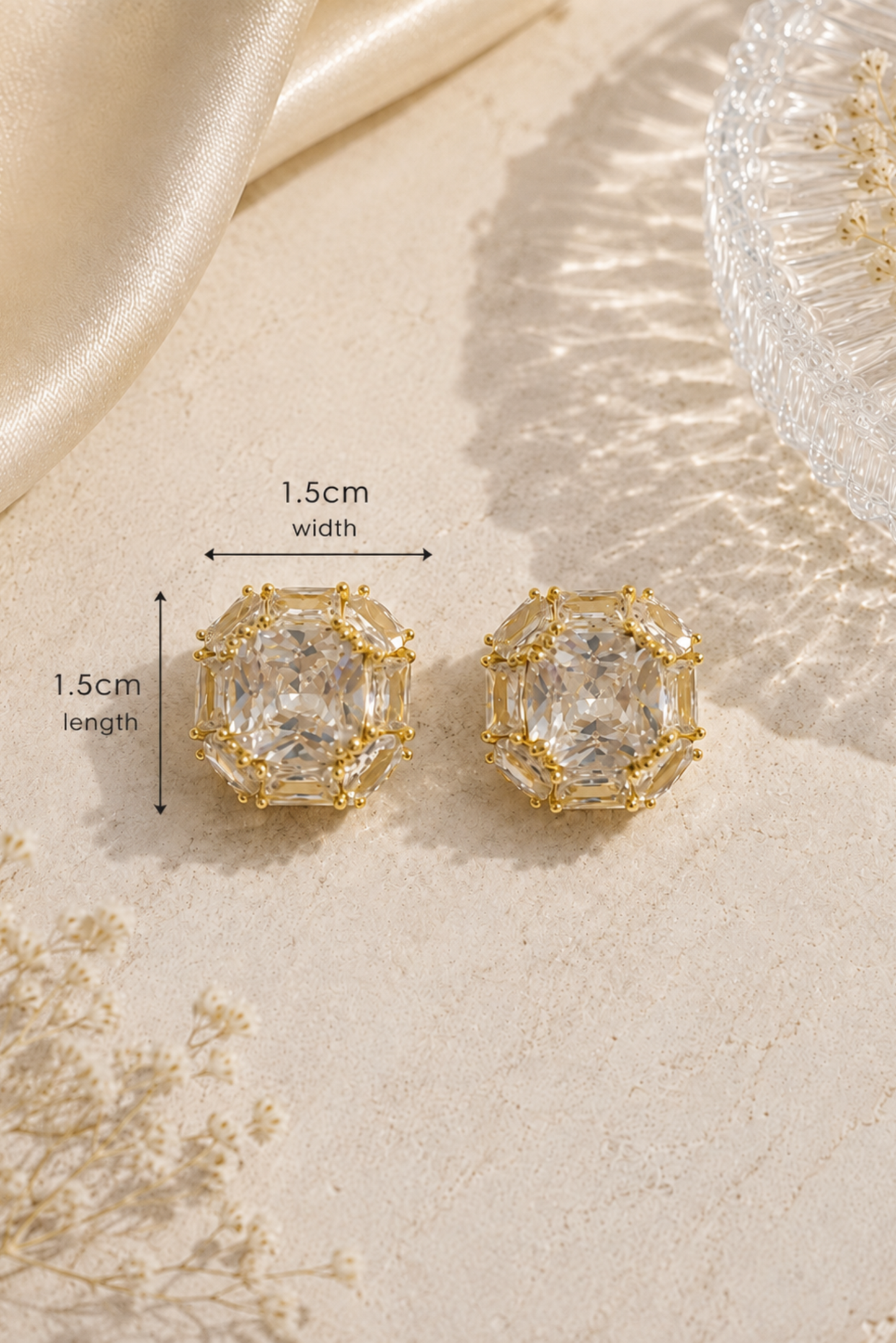 A pair of gold-toned stud earrings with large clear rectangular gemstones, shown on a light beige textured surface with satin fabric, dried flowers, and a glass dish. Black measurement arrows indicate 1.