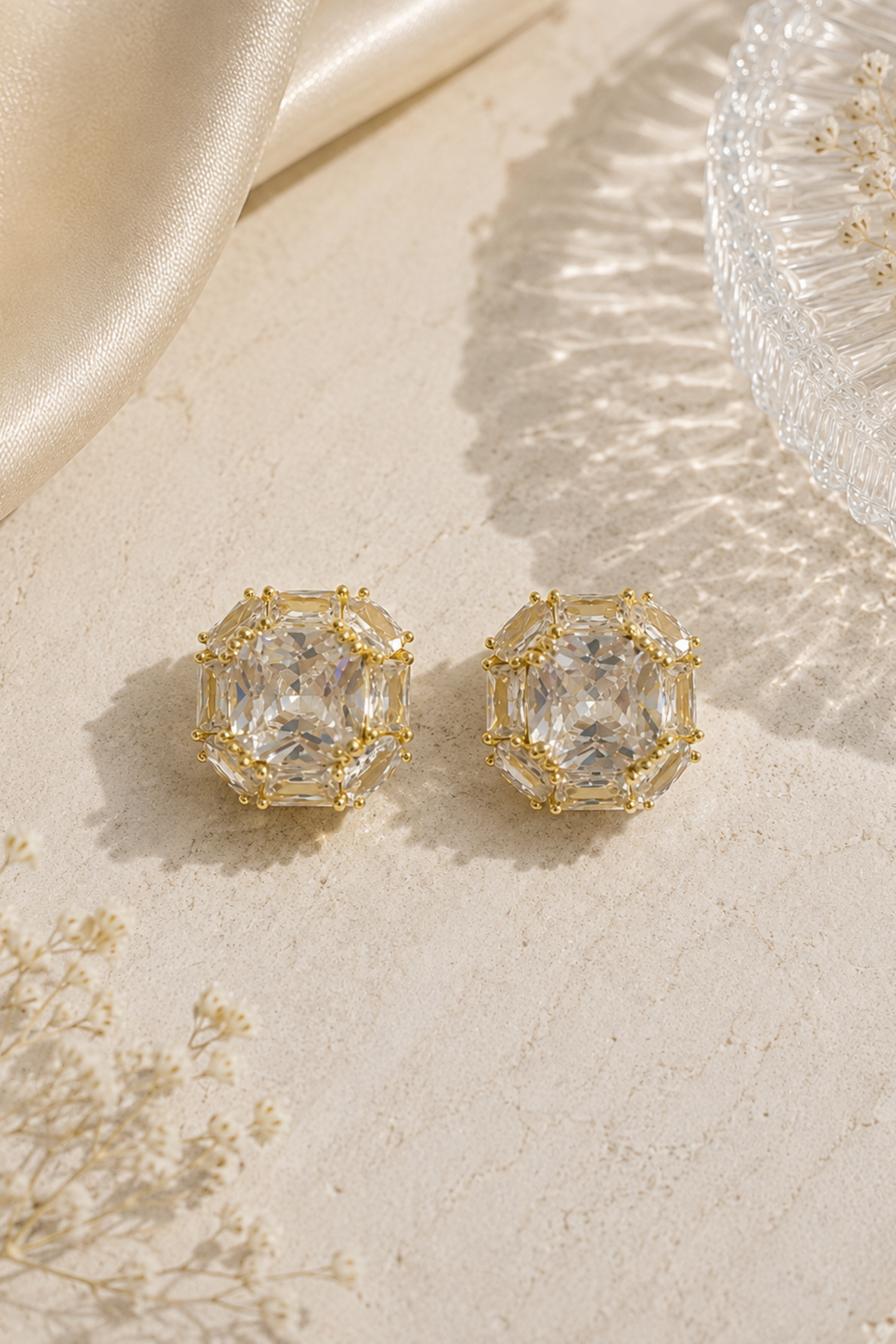 A pair of gold-toned stud earrings with large clear rectangular gemstones, styled on a textured cream surface with satin fabric and dried flowers.