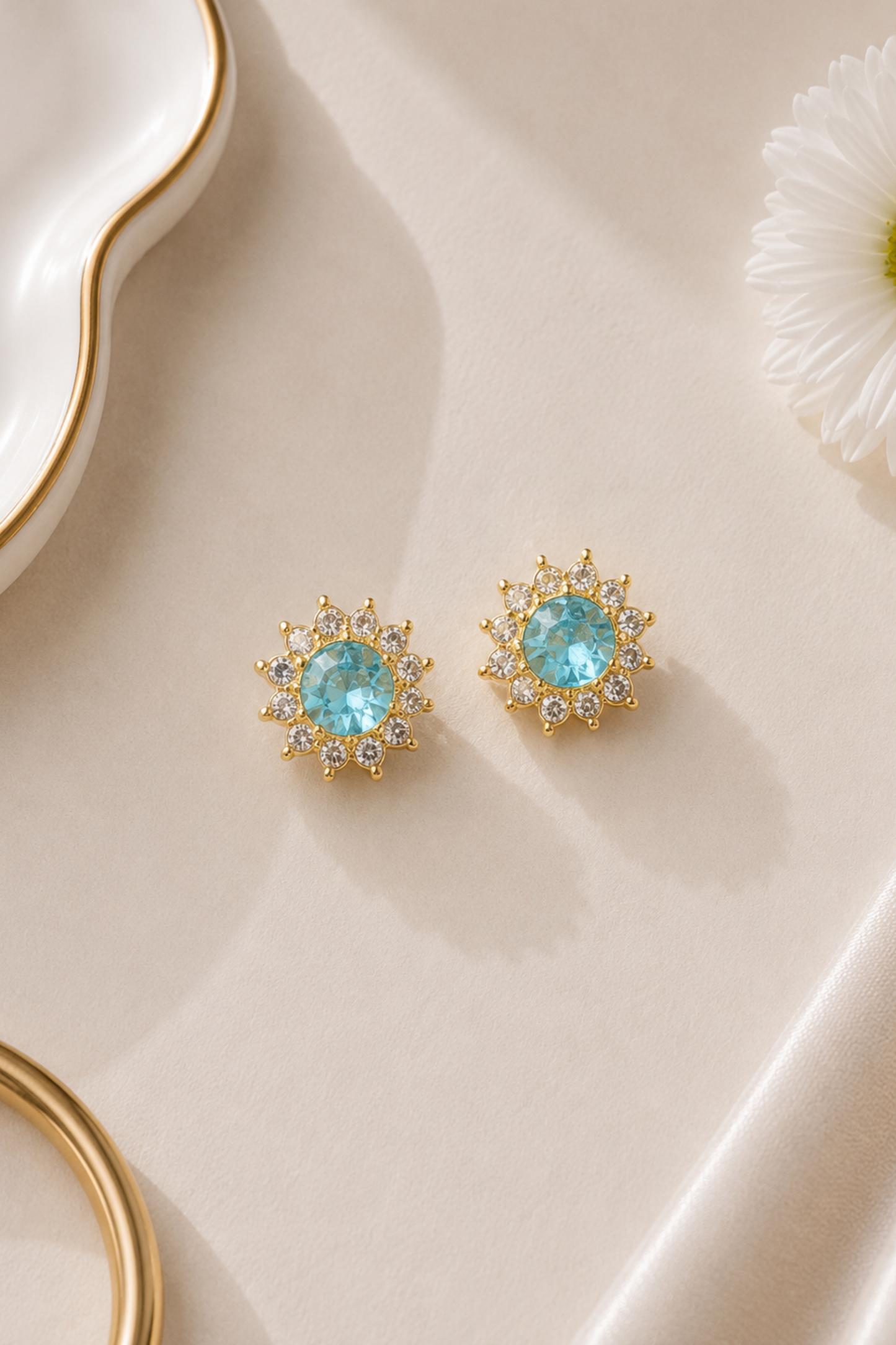 A pair of gold-toned stud earrings with round light blue center stones and small clear surrounding stones, arranged on a warm cream surface with decorative props.