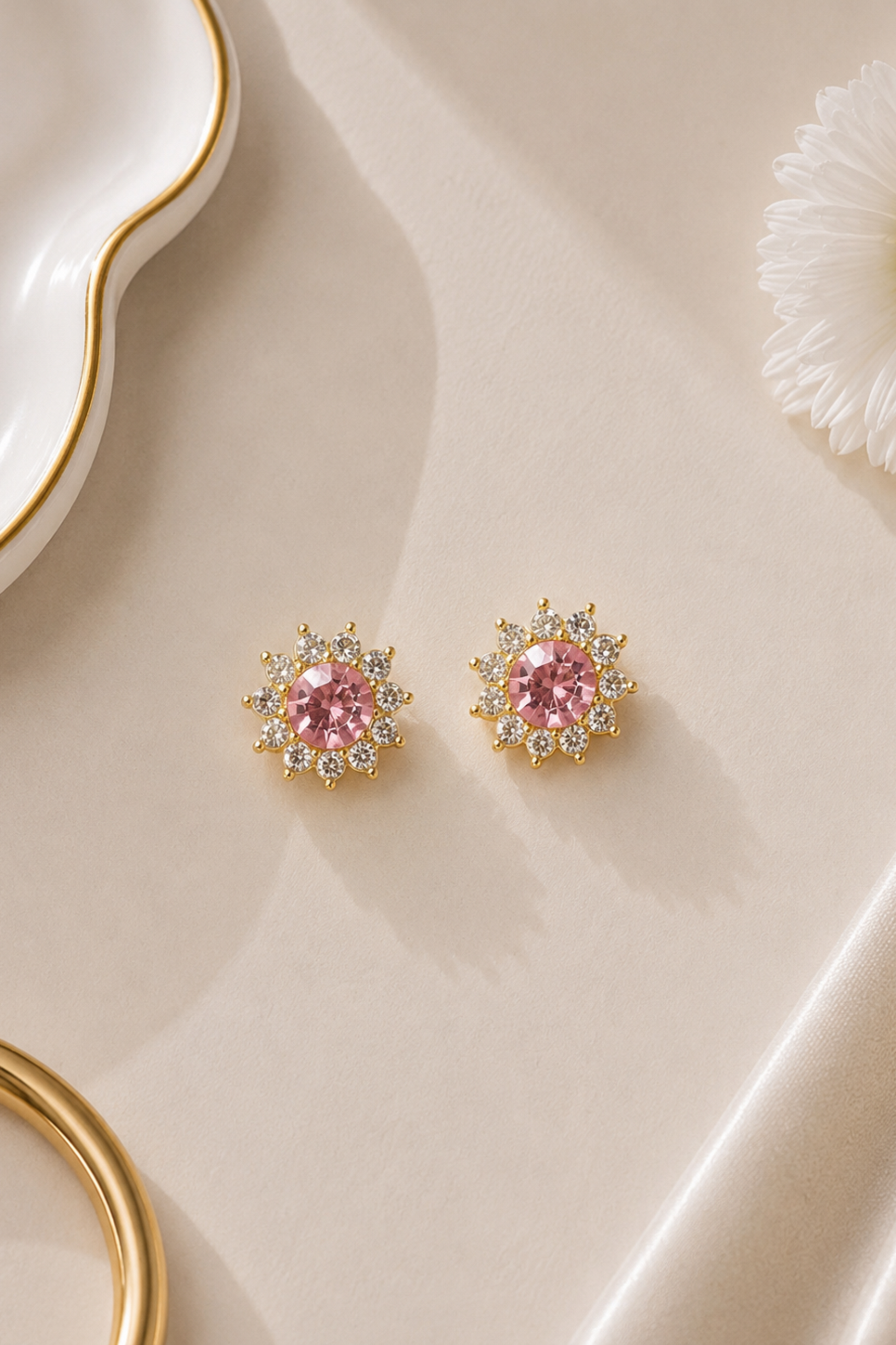 A pair of gold-toned stud earrings with round pink center stones and small clear surrounding stones, arranged on a soft cream surface with decorative props.