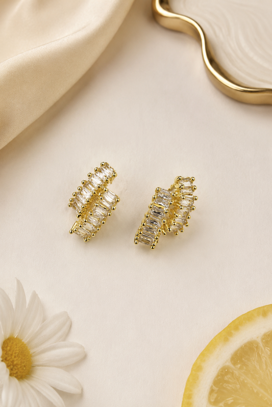 A pair of gold-toned wrap-style earrings with clear rectangular stones, arranged on a light cream surface with a cream fabric fold, a white daisy flower, and a lemon slice.