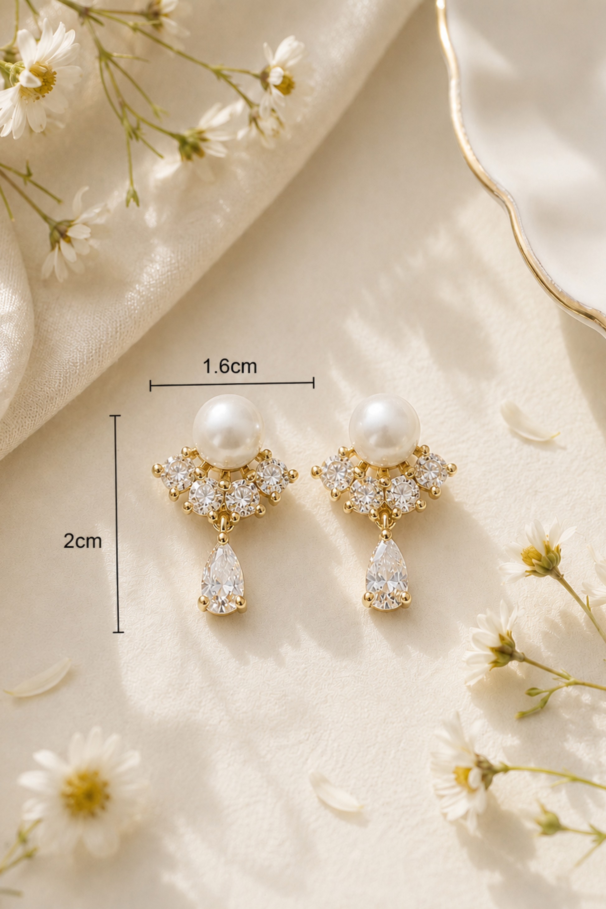 A pair of gold and pearl drop earrings displayed on a cream satin fabric with daisy flowers. Black measurement lines and text indicate the dimensions: 1.
