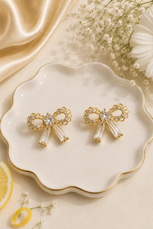 A pair of gold bow-shaped stud earrings with clear rhinestone accents and clear crystal ribbon tails, displayed on a scalloped white ceramic dish with gold trim, surrounded by decorative flowers, baby's breath, a lemon slice, and draped satin fabric.