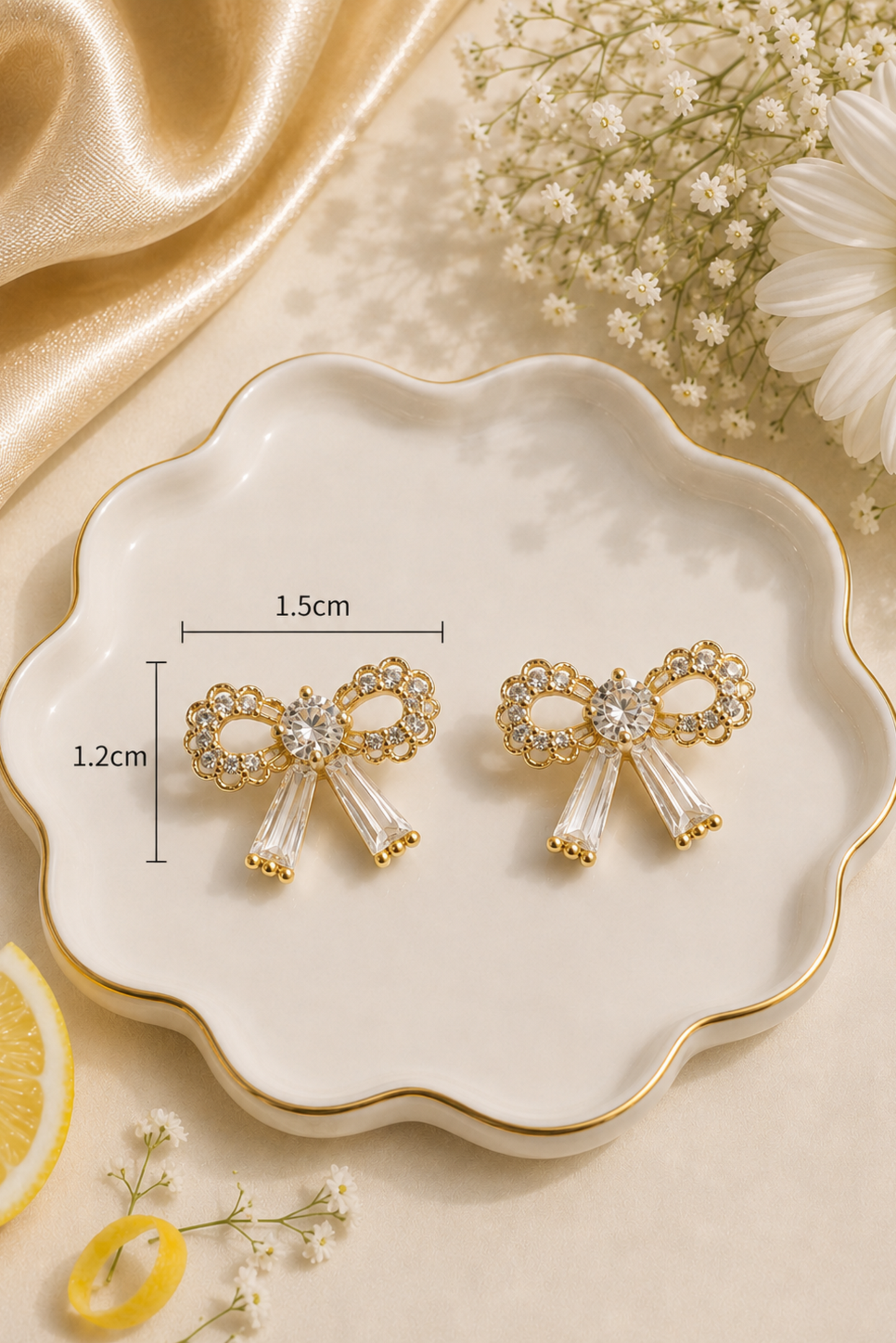 A pair of gold bow-shaped stud earrings with clear rhinestone accents and dangling clear crystal strands, displayed on a scalloped white dish with measurement labels showing 1.5 cm width and 1.