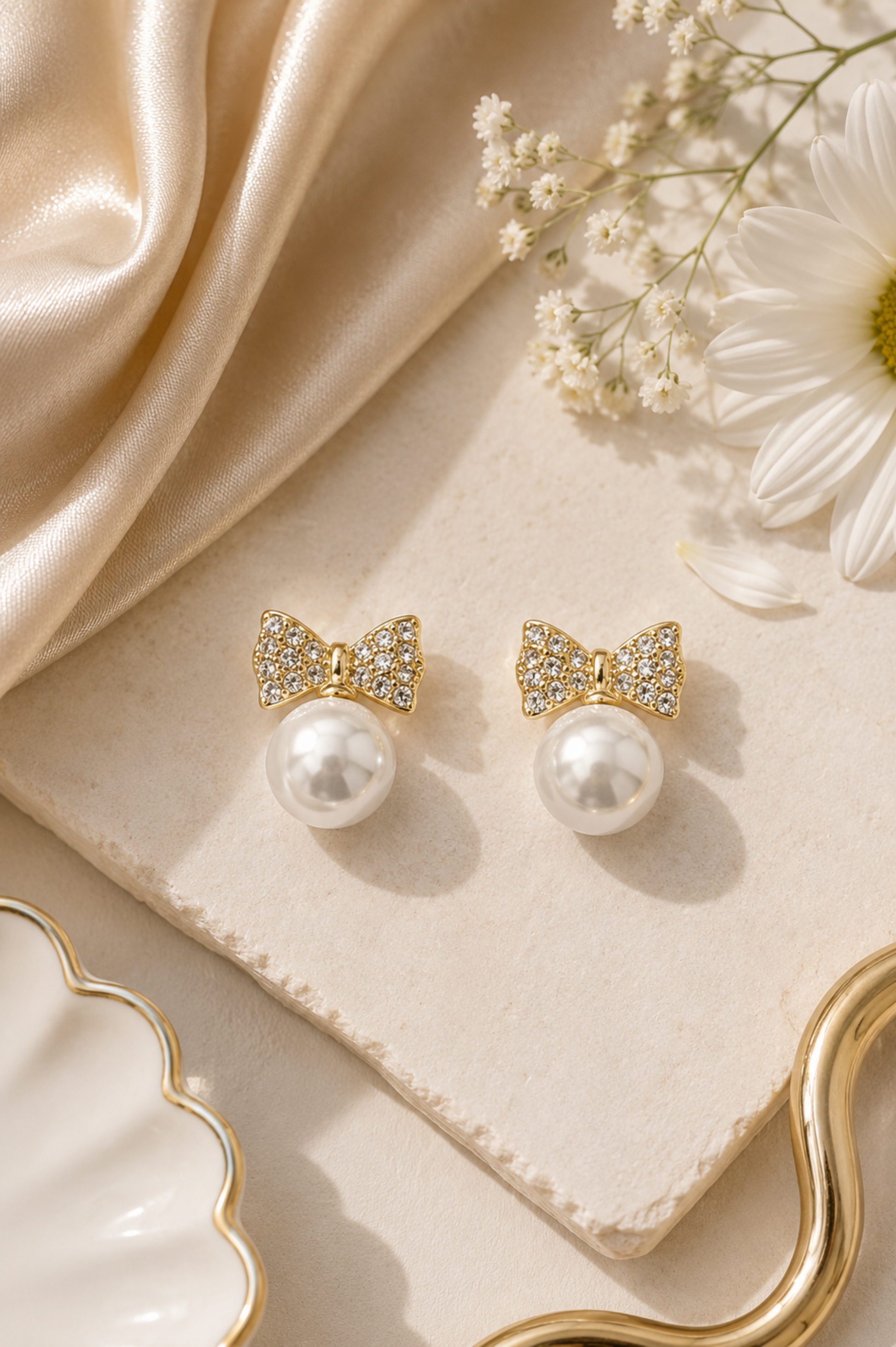 A pair of gold bow stud earrings with clear crystal detailing and white pearl drops, styled on a beige stone slab with cream satin fabric, white flowers, and a scalloped dish.
