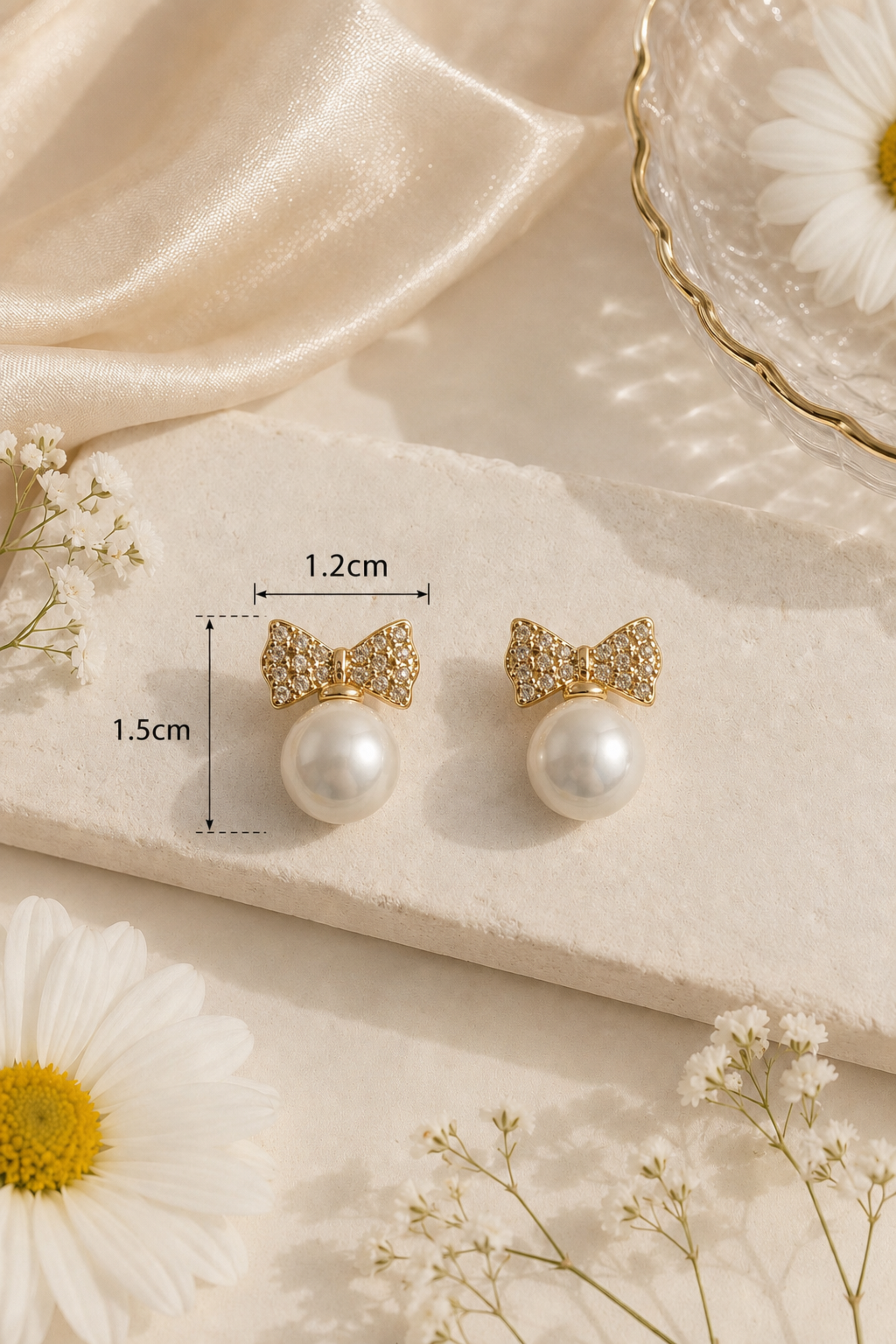 A pair of gold bow stud earrings with clear rhinestones and white pearl drops, displayed on a beige stone slab with dimension lines showing 1.2cm width and 1.