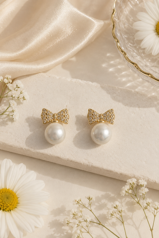 A pair of gold bow stud earrings with sparkling crystal-like detailing and a white pearl drop, styled on a cream stone slab with white daisies and satin fabric.