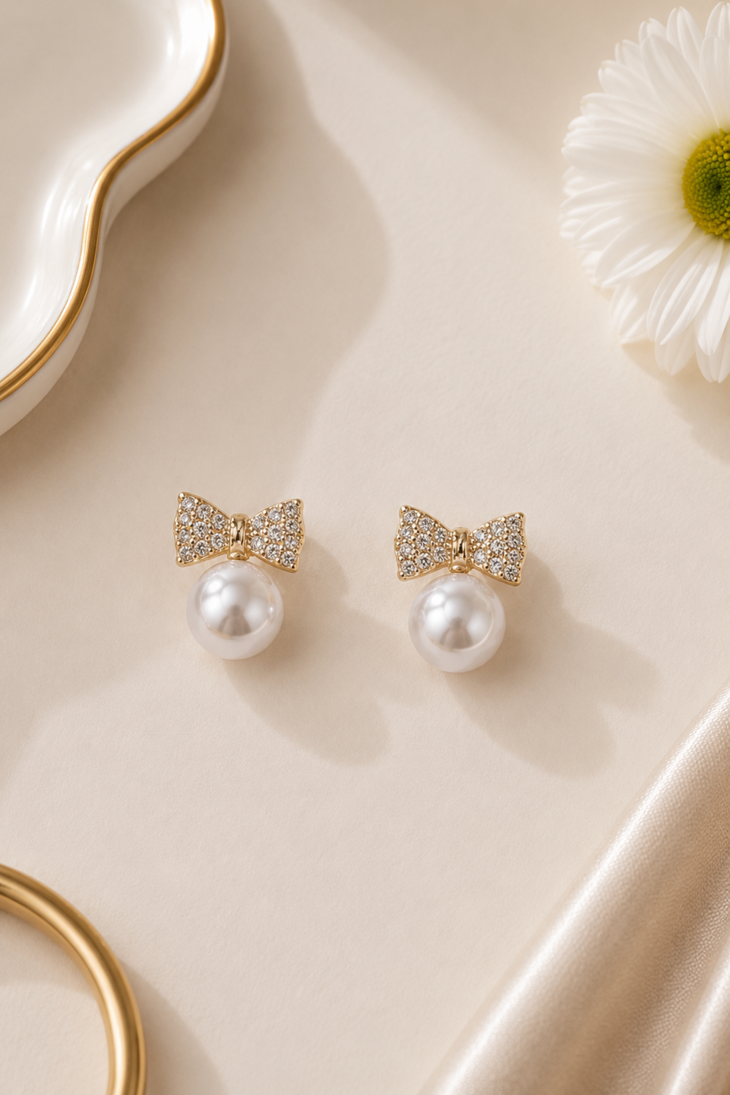 A pair of gold bow stud earrings with white pearl drops and sparkling crystal details, displayed on a beige surface with a white daisy flower and gold-toned props.