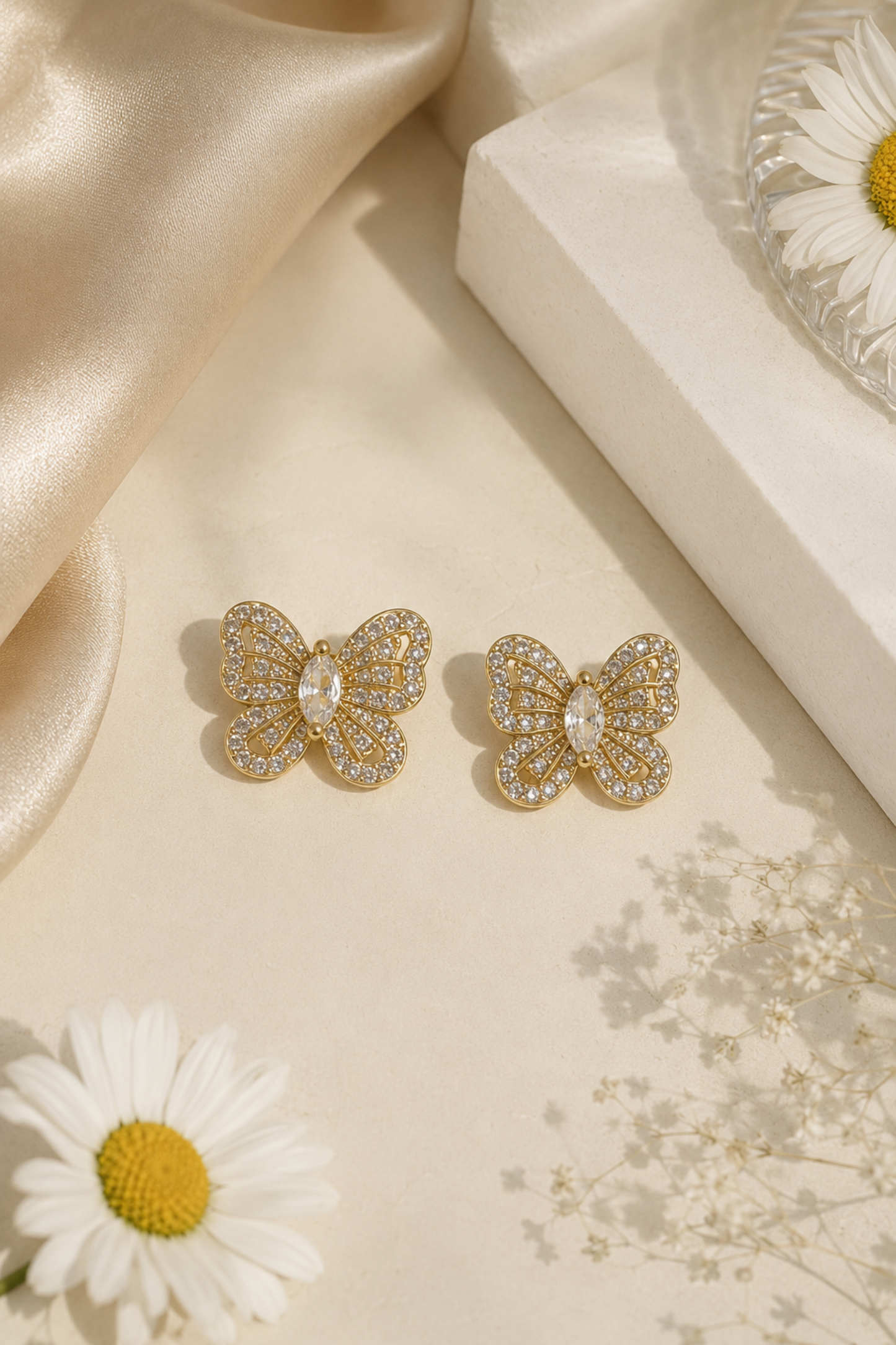 A pair of gold butterfly stud earrings adorned with clear crystals, displayed on a beige surface with a cream gift box, satin fabric, and white daisies.
