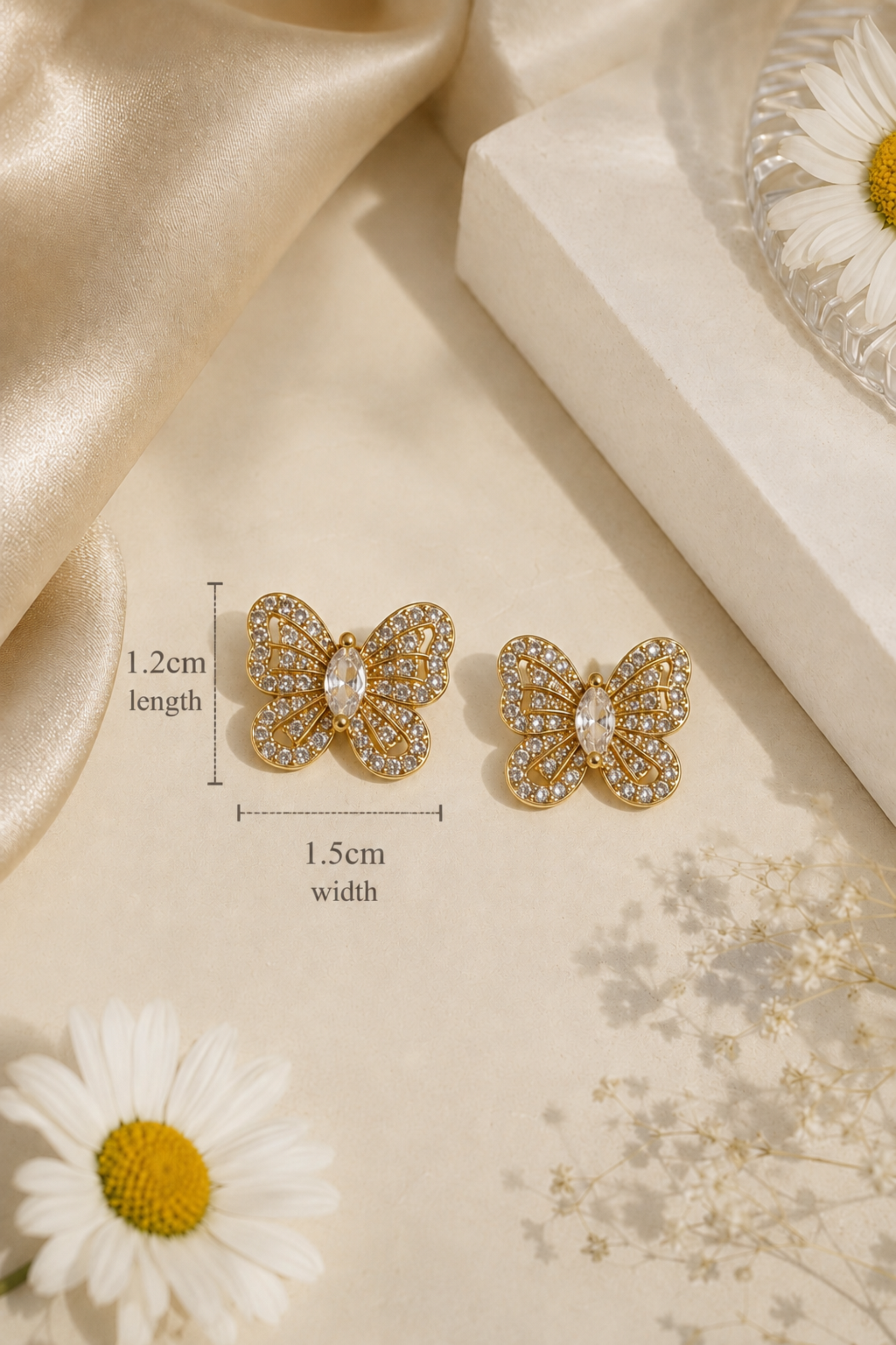 A pair of gold butterfly stud earrings with clear rhinestones, displayed on a beige surface with daisy flowers and satin fabric. Measurement lines and text indicate the earrings are 1.