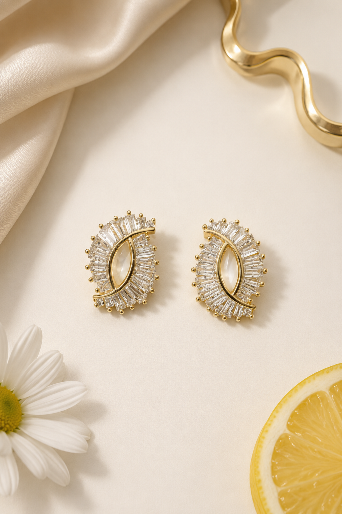 A pair of gold crescent-shaped stud earrings adorned with clear baguette and round crystals, styled on a cream background with a white daisy, a lemon slice, and draped satin fabric.
