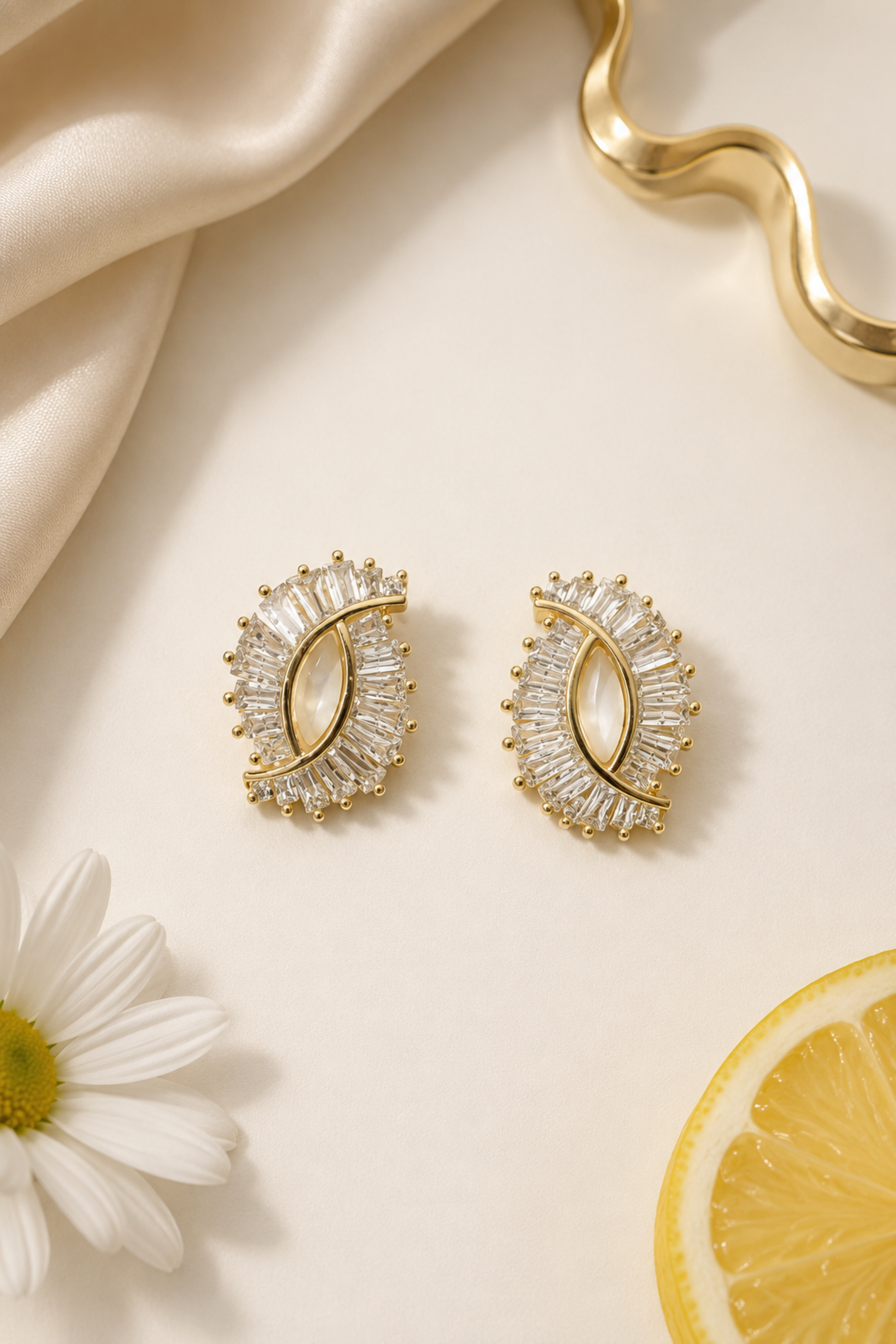 A pair of gold crescent-shaped stud earrings adorned with clear baguette and round crystals, styled on a cream background with a white daisy, a lemon slice, and draped satin fabric.