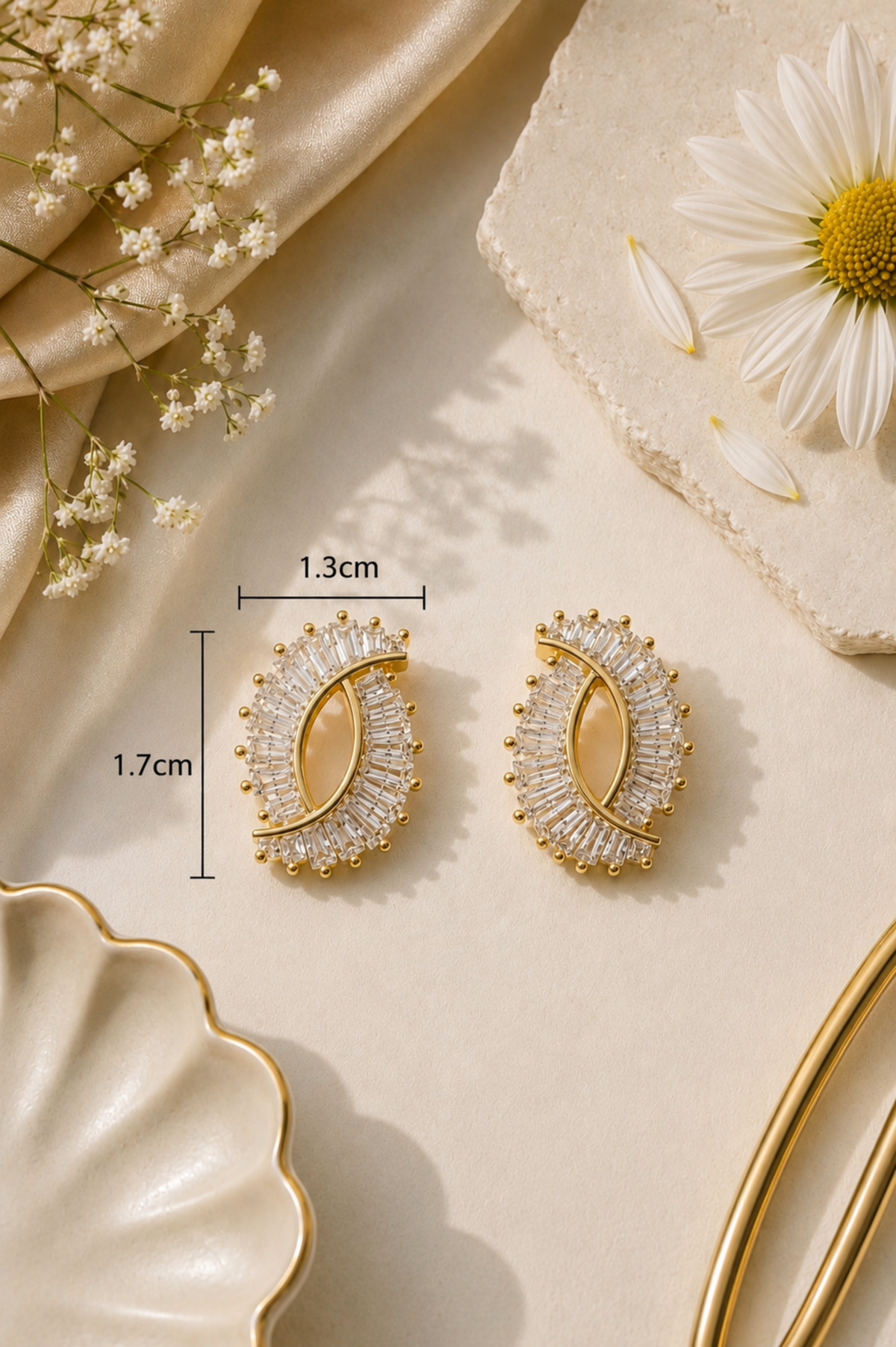 A pair of gold crescent-shaped stud earrings with clear crystal-like stones and a central gold oval, shown on a beige surface with decorative flowers and fabric. Black measurement lines and text indicate 1.