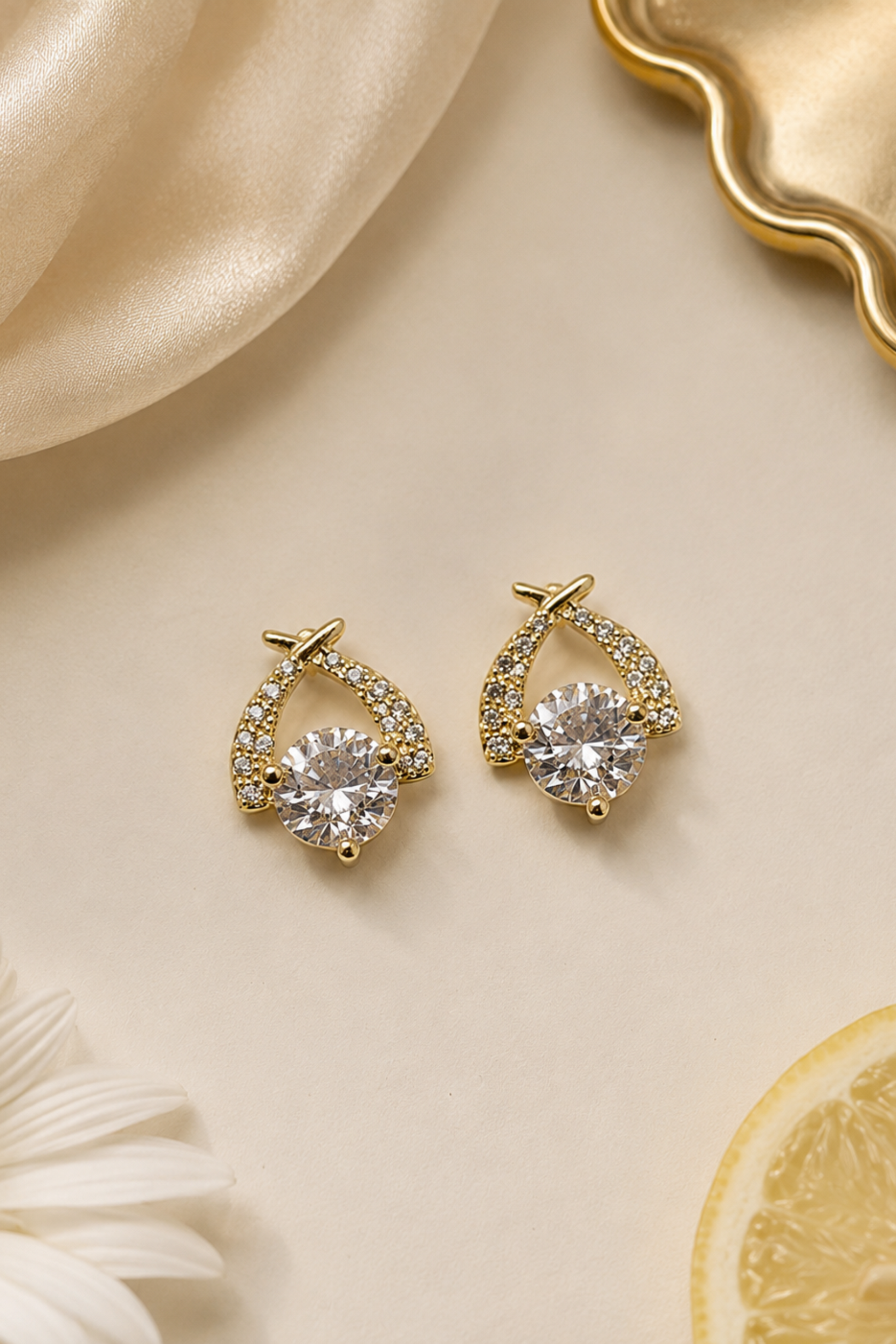 A pair of gold crescent-shaped stud earrings with clear round-cut center stones and small pavé-set accent stones, arranged on a warm cream surface with satin fabric, a daisy flower, and a lemon slice.