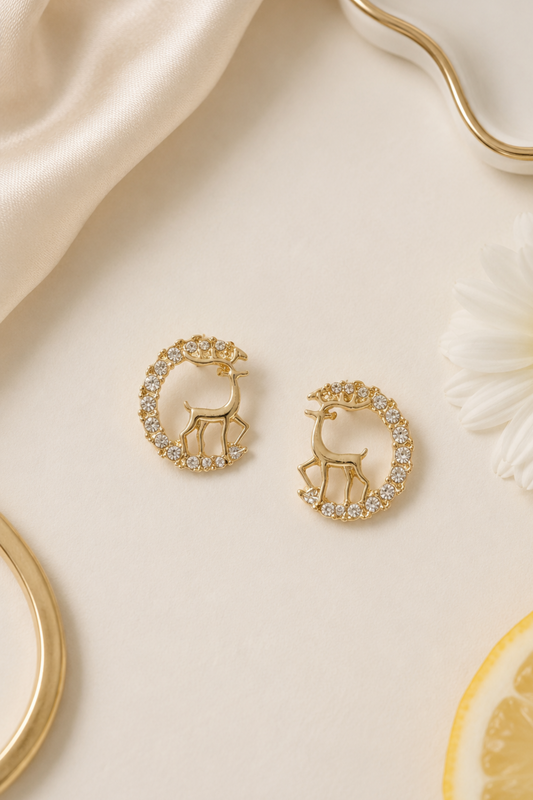 A pair of gold crescent moon and deer stud earrings decorated with clear crystals, displayed on a warm cream surface with a satin fabric, a daisy flower, and a lemon slice.