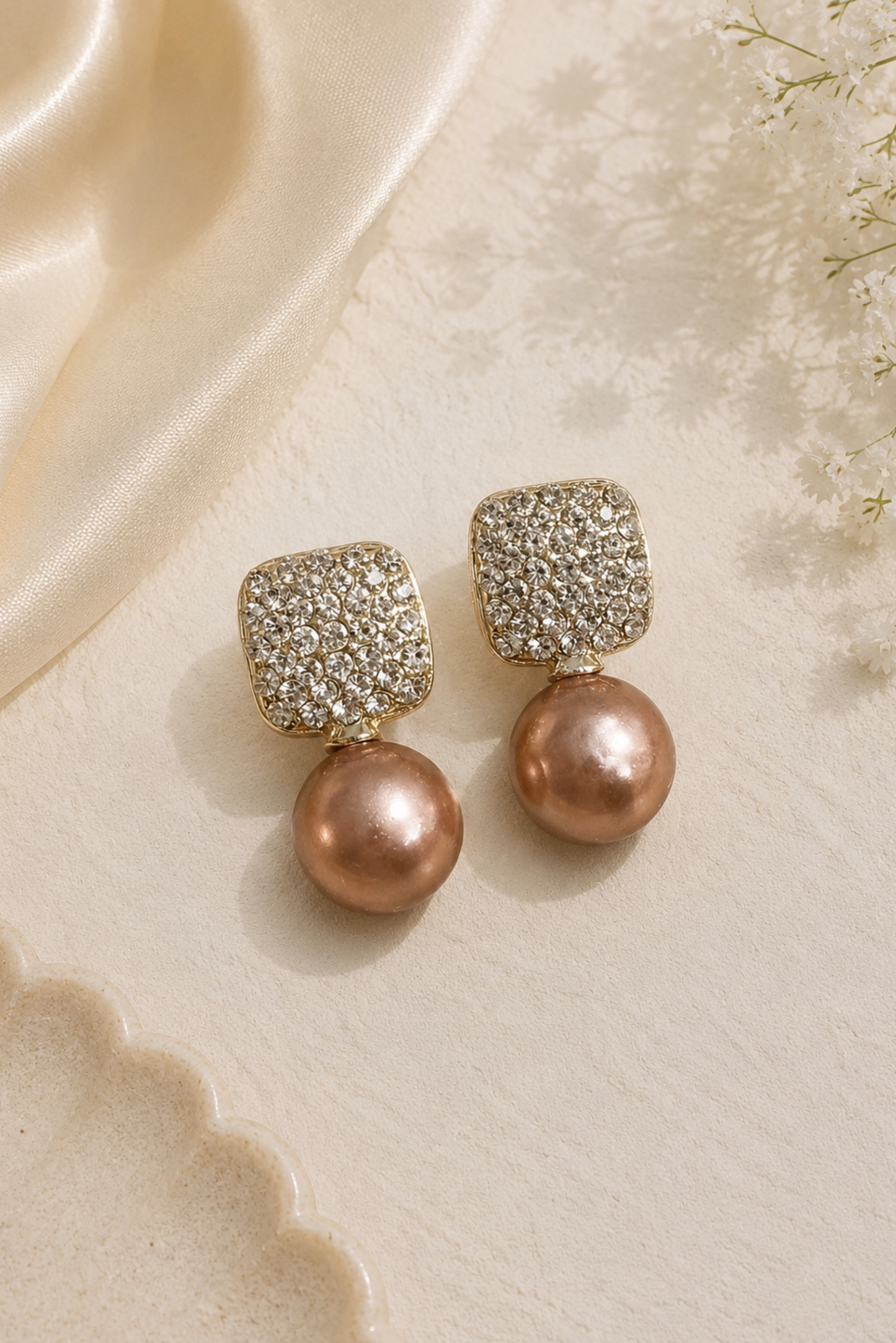 A pair of gold drop earrings featuring a square top covered in clear pavé stones and a dangling round rose gold pearl, displayed on a textured cream surface with a cream satin ribbon.