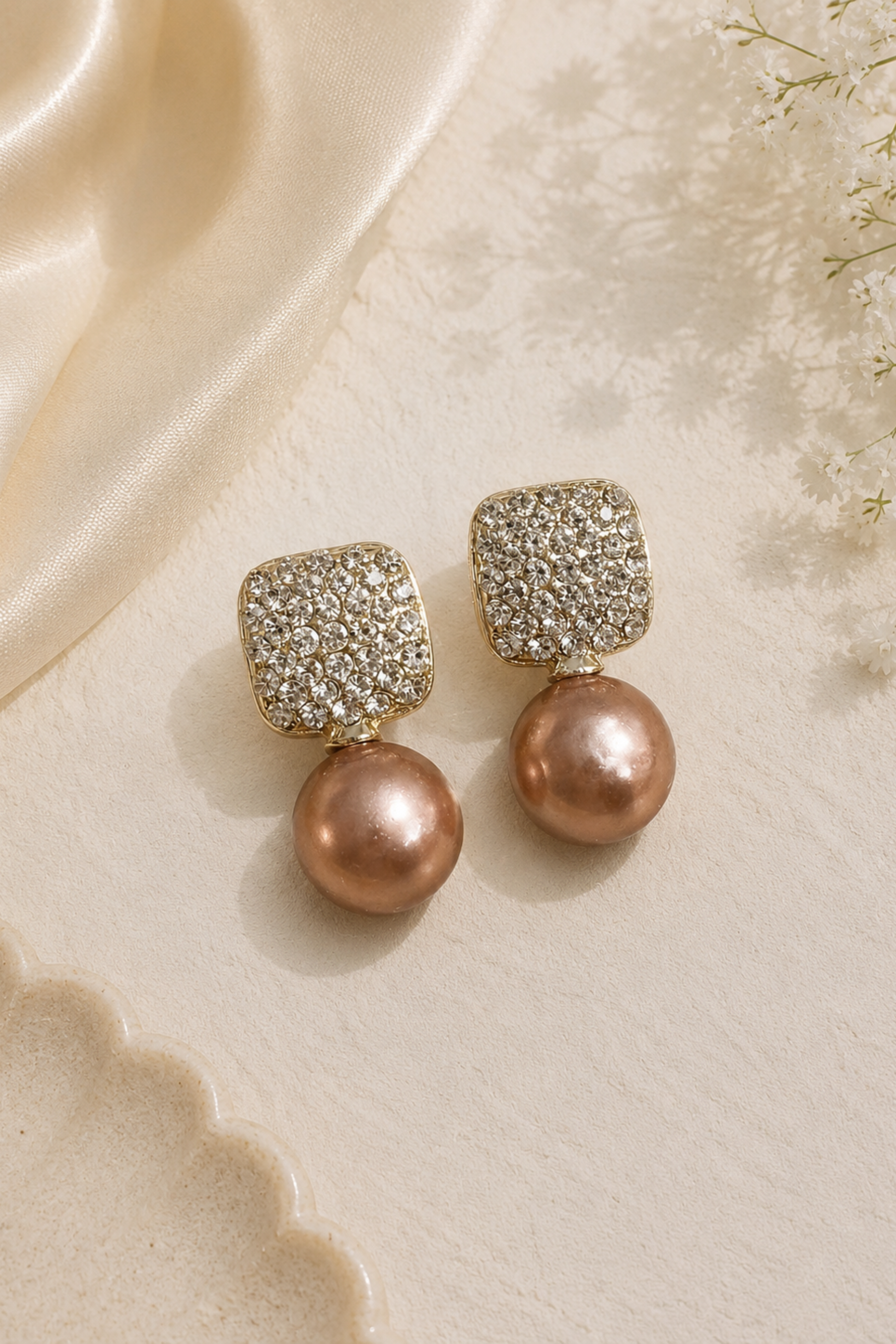 A pair of gold drop earrings featuring a square top covered in clear pavé stones and a dangling round rose gold pearl, displayed on a textured cream surface with a cream satin ribbon.