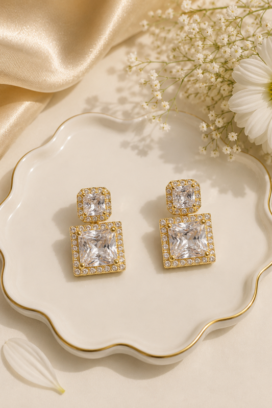 A pair of gold drop earrings with clear rectangular gemstones, displayed on a scalloped cream dish with gold trim, surrounded by white baby's breath flowers and a gold satin fabric.