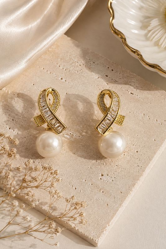 A pair of gold drop earrings with clear rectangular stones and a large white pearl dangling below, styled on a beige stone slab with satin fabric and dried flowers.