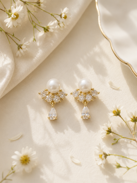 A pair of gold drop earrings with white pearls and clear crystal accents, styled on a cream satin fabric with white daisies.
